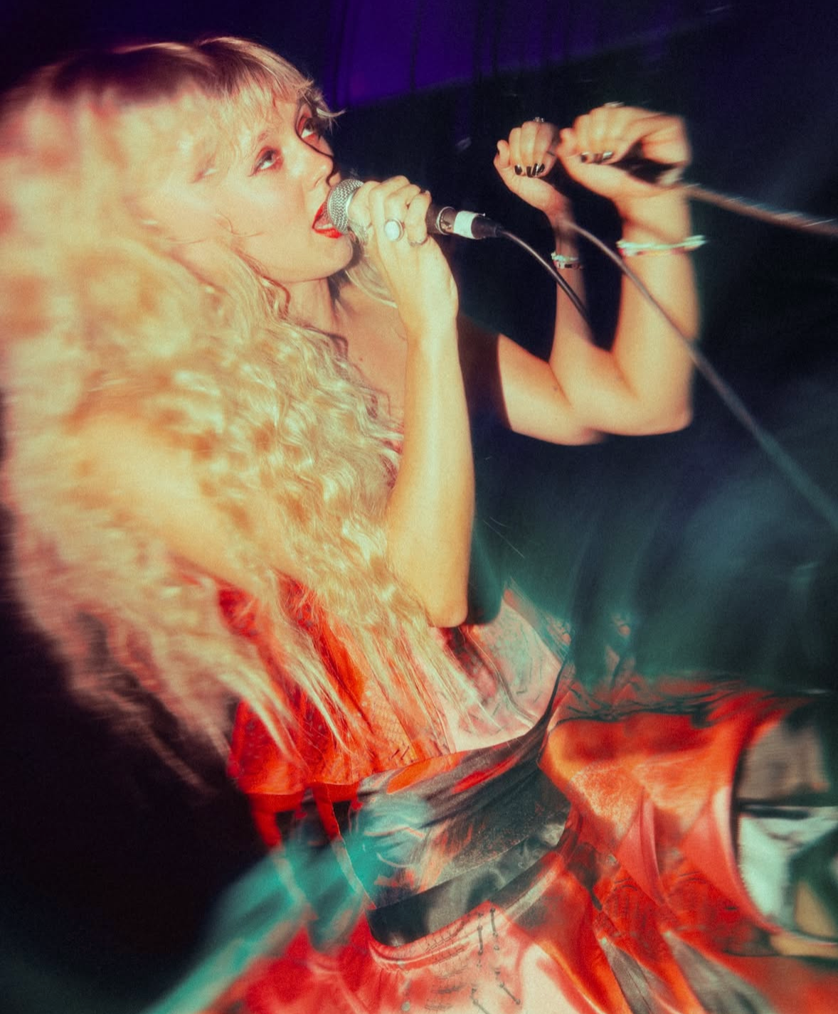 A woman with long, curly blonde hair singing into a microphone, with her eyes closed and a passionate expression. She is wearing a sleeveless top and bracelets, with a dark background and colorful lighting.