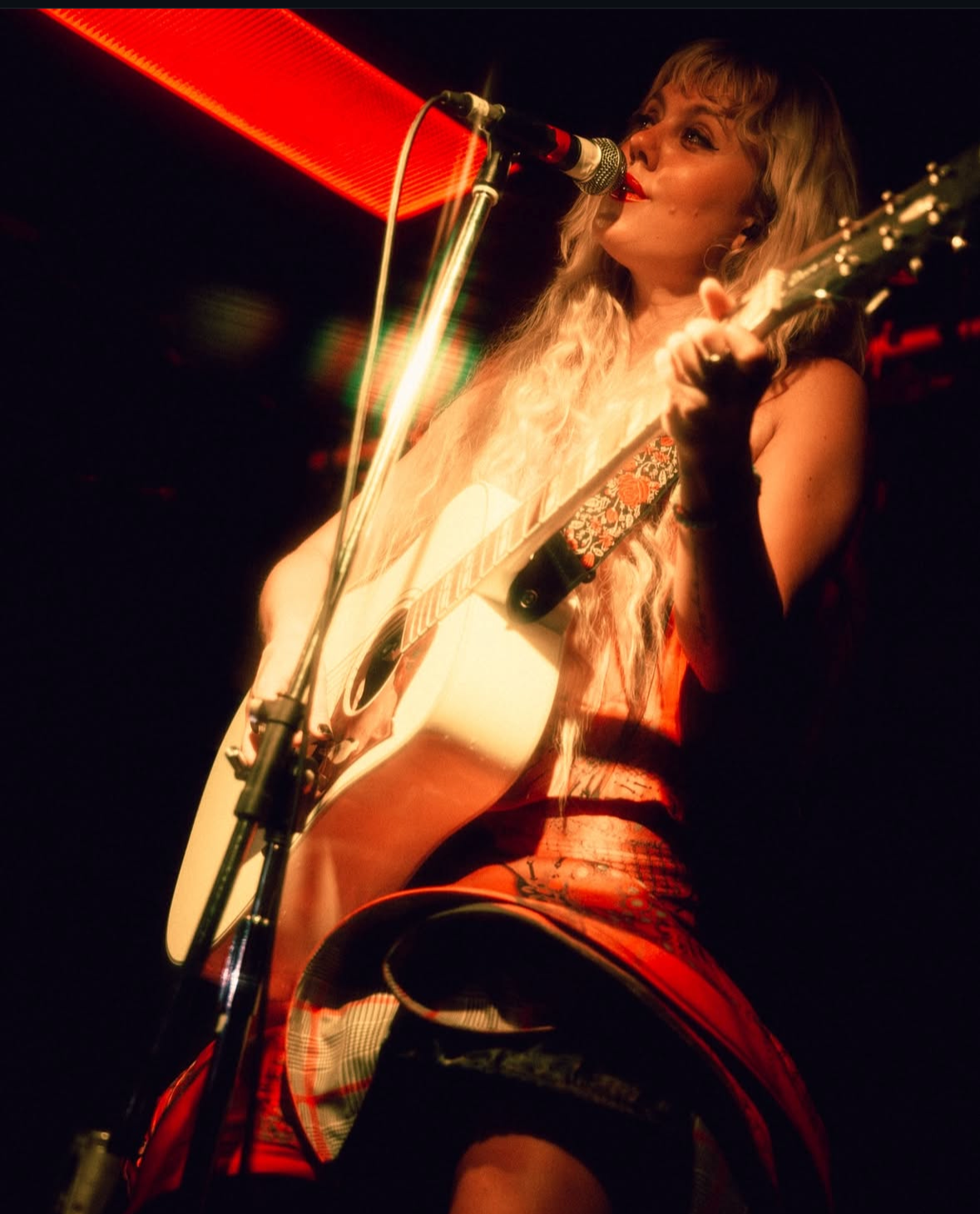 Female musician performing with guitar and singing into a microphone on stage with red lighting.