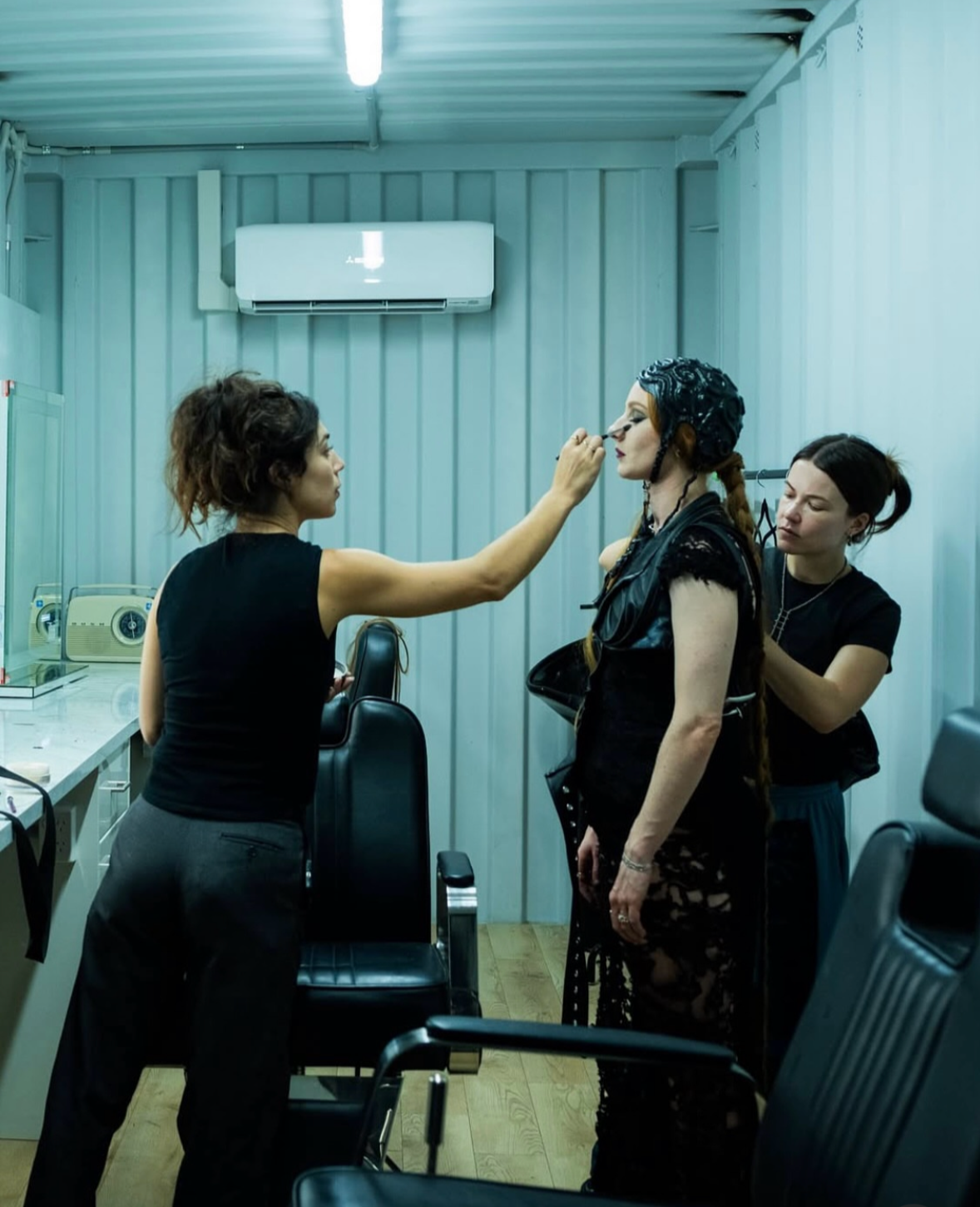 A woman applying makeup to another woman in a backstage setting with makeup tools and chairs around.
