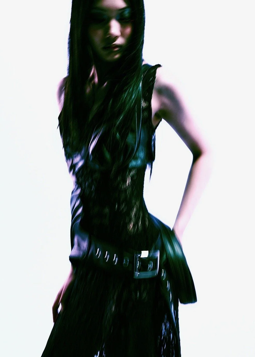 A woman with long dark hair wearing a black leather outfit and belt, posing against a plain white background.