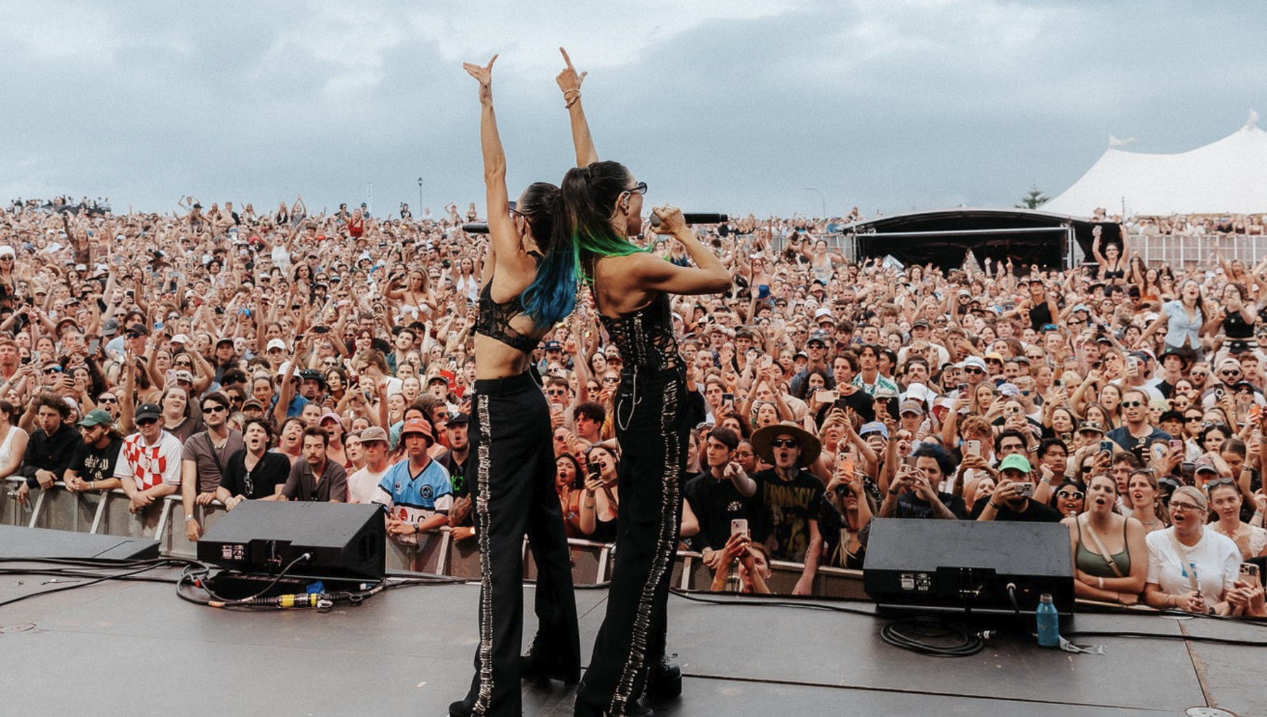 Two female performers with rainbow-colored hair singing on stage in front of a large outdoor crowd at a concert.