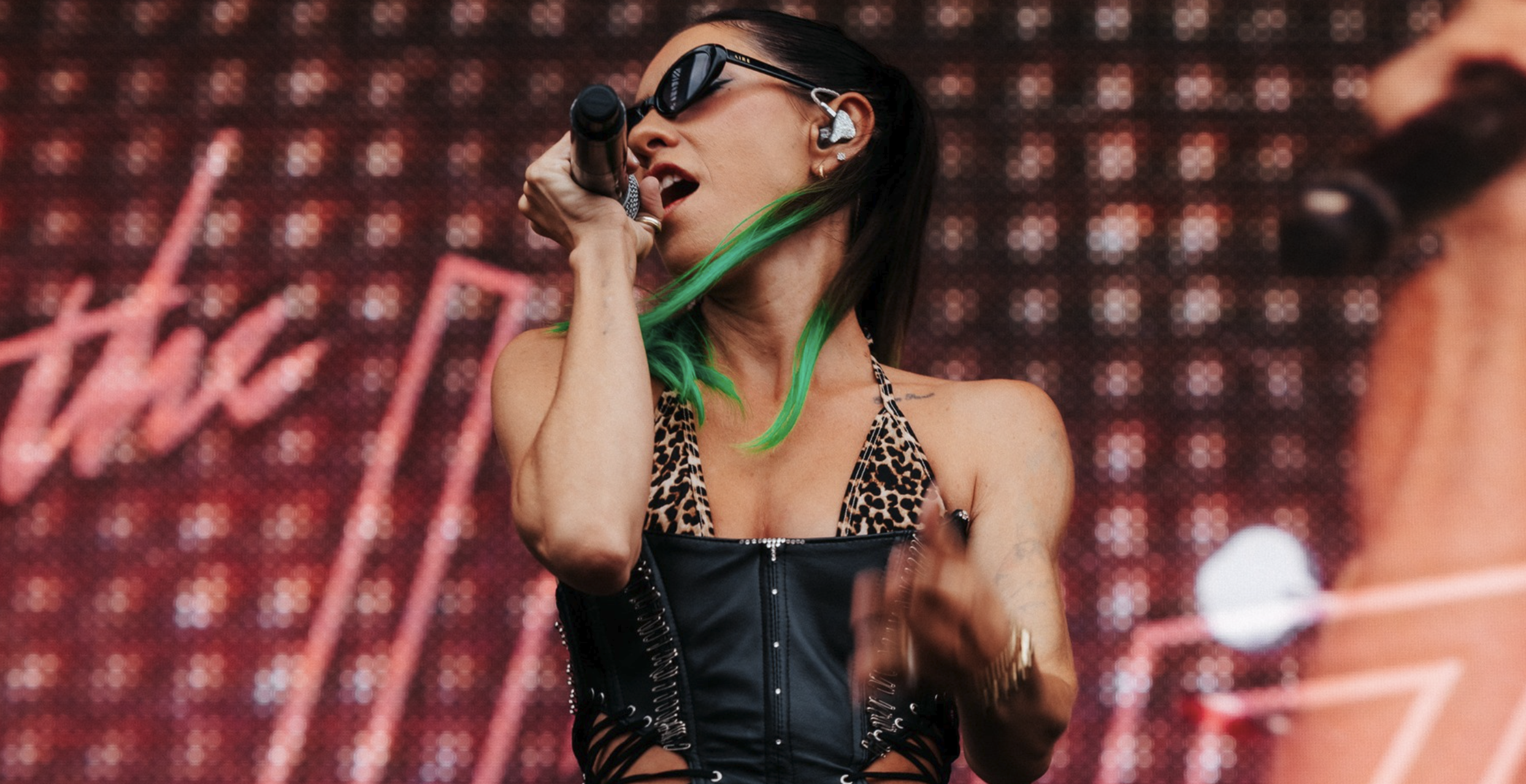 A woman singing into a microphone on stage, wearing sunglasses, leopard print top, and a black leather corset, with green hair and in-ear monitors, with a blurred background featuring red and white lights.
