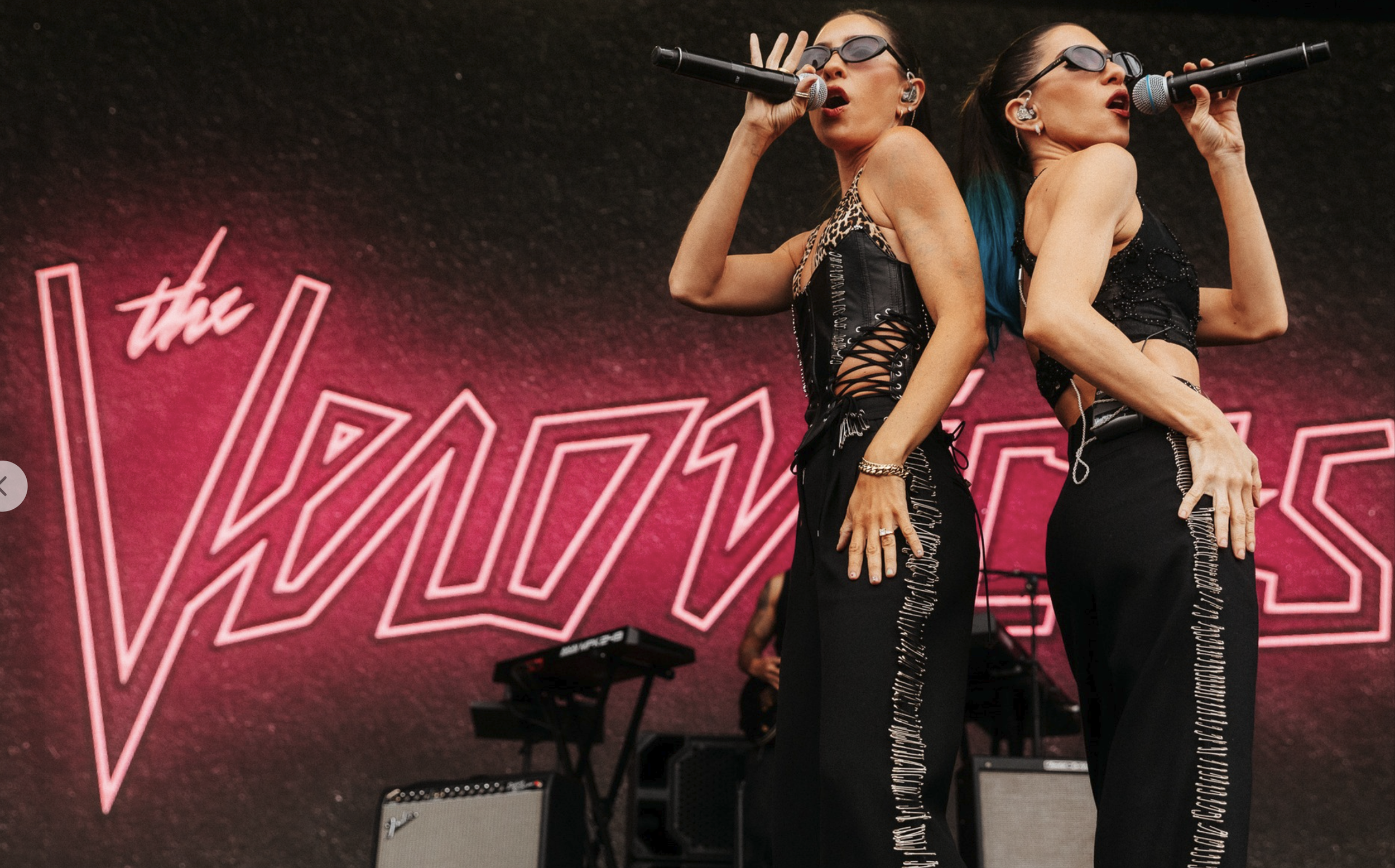 Two women with long dark hair and black sunglasses singing into microphones on stage, wearing black outfits with silver details, with a pink neon sign in the background.