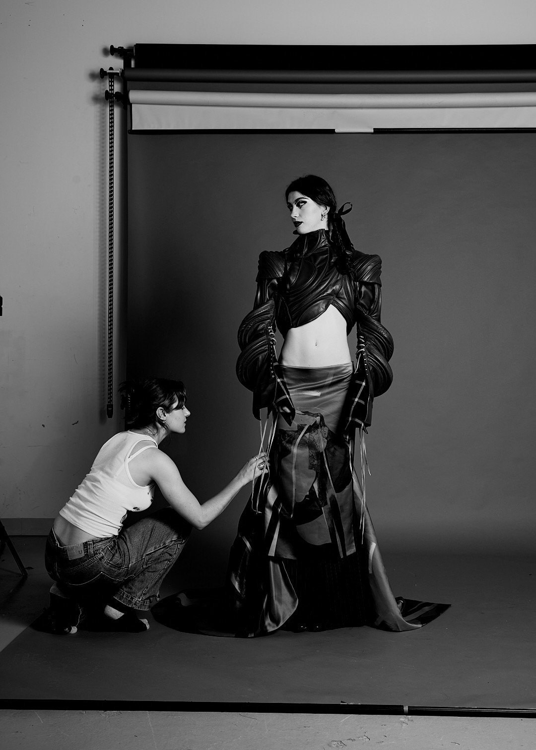 A woman with dark hair and makeup wearing an edgy, futuristic leather outfit poses in front of a gray backdrop during a photoshoot. A photographer kneels on the floor adjusting her clothing.