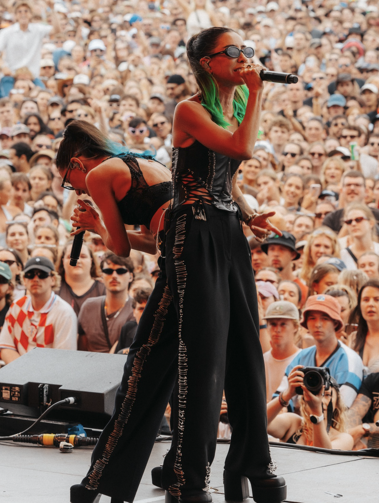 A woman with green hair and wearing sunglasses, dressed in a black corset top and black wide-leg pants, singing into a microphone on stage at a crowded outdoor concert. The audience is diverse, wearing casual clothing and sunglasses, with some taking