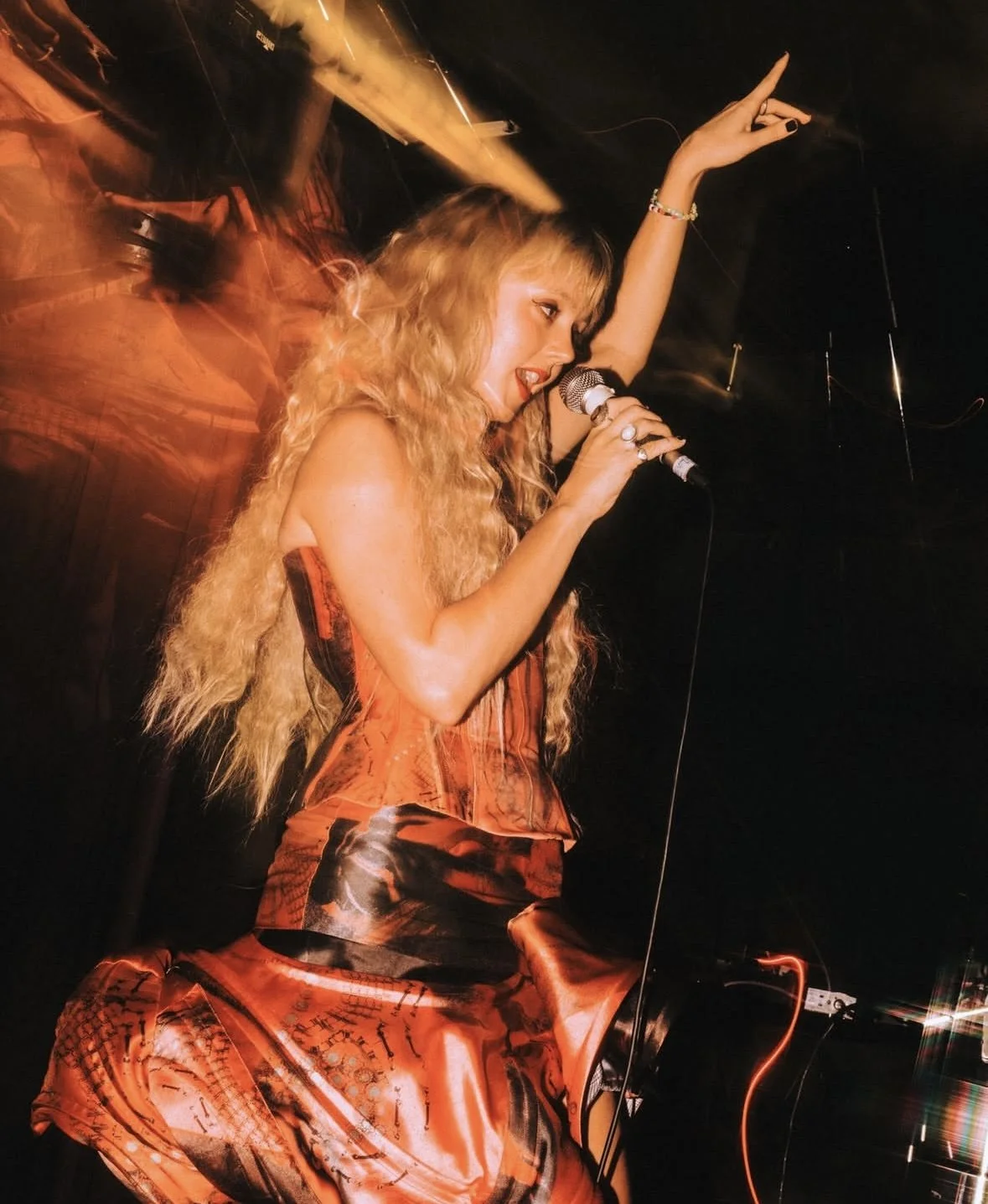 A woman with long, curly blonde hair singing into a microphone on stage, wearing an orange and black patterned dress, with arms raised and a dark background.