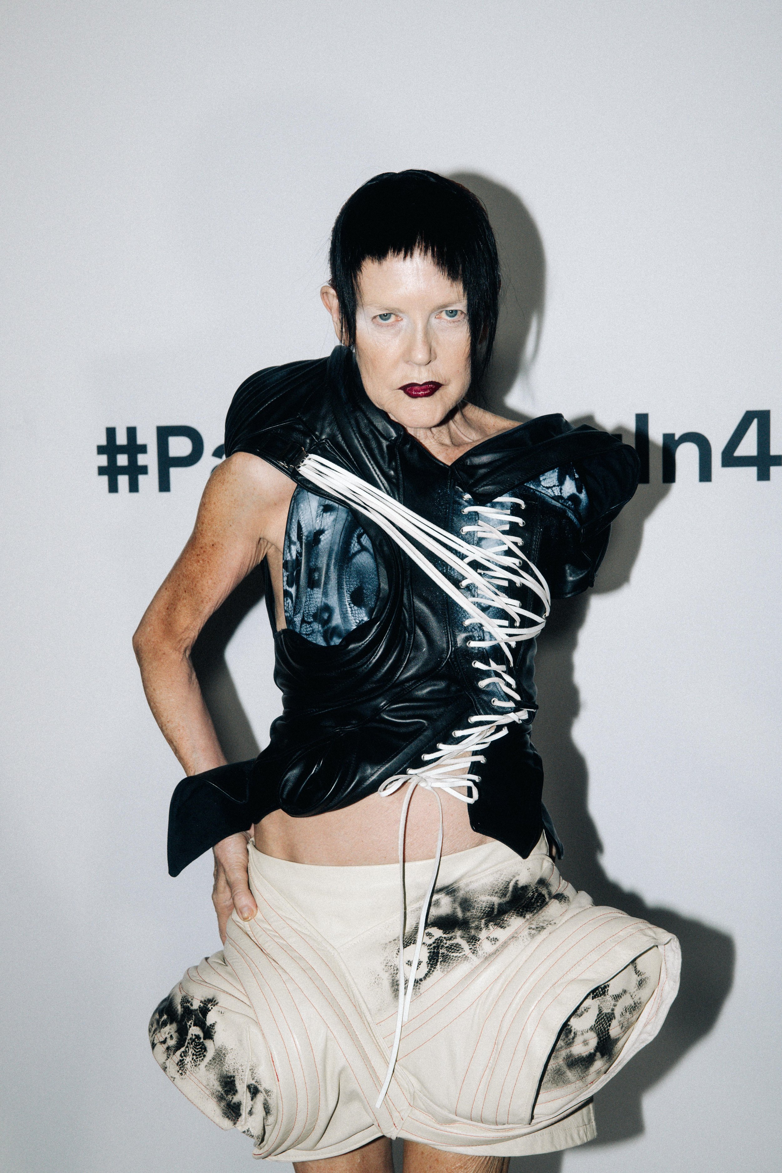 An androgynous model with short black hair, piercing blue eyes, and pale skin, wearing a black leather vest with white lacing, beige shorts, and patterned pants, posing against a white background with hashtags.