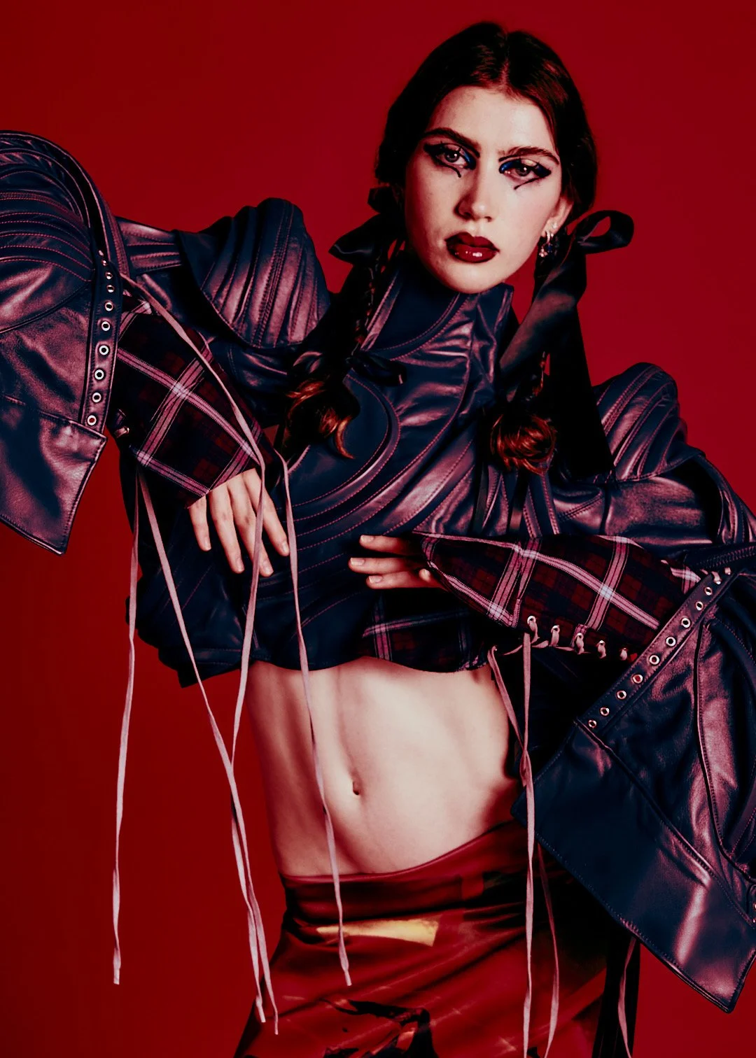 A woman with dark hair and makeup wearing a black leather jacket with plaid sleeves, revealing her midriff, against a red background.