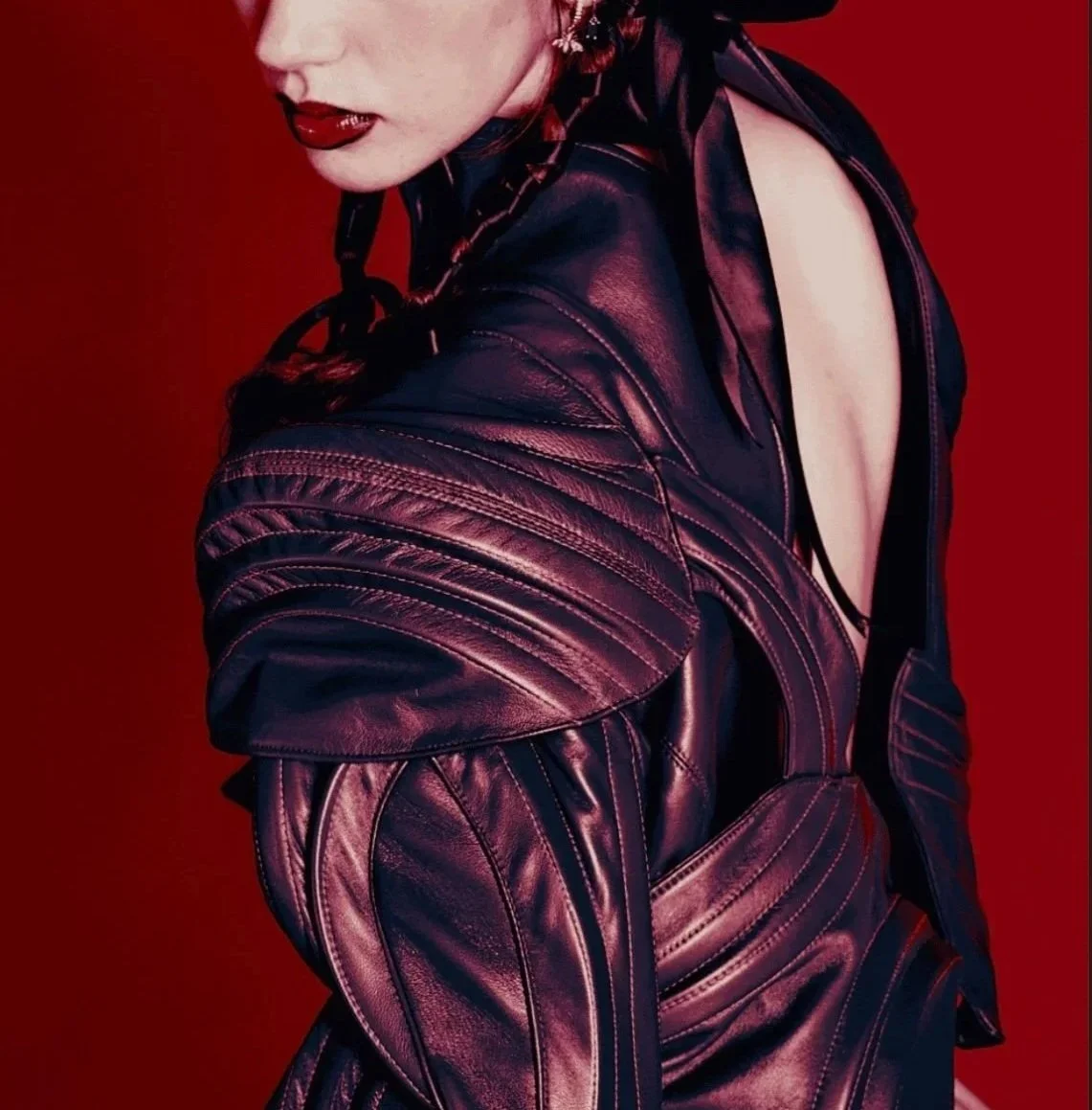 Close-up of a woman wearing a shiny black leather jacket, with her face slightly turned and lips painted red, against a red background.