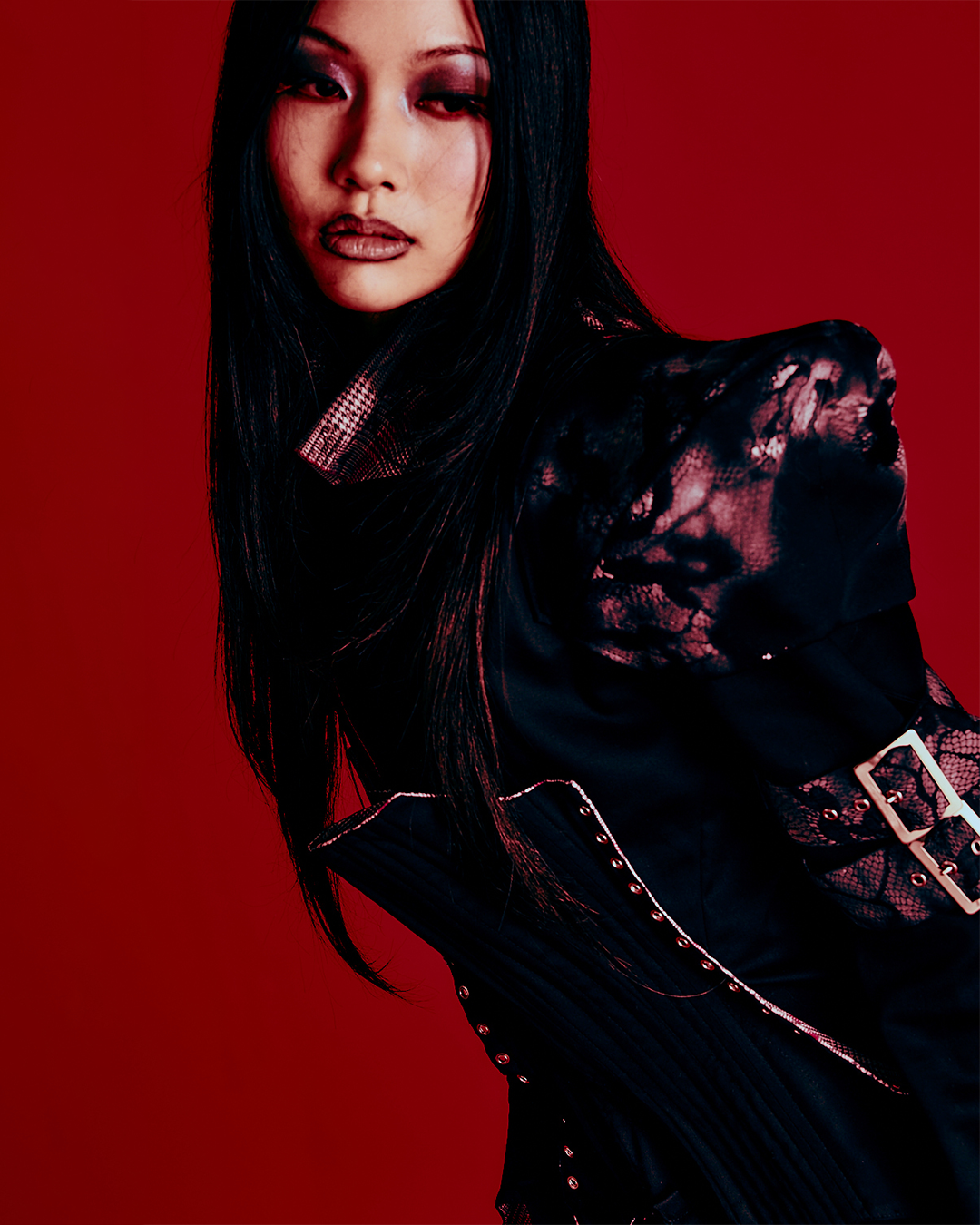 A woman with long black hair, wearing dark makeup, a black patterned jacket with lace details and buckles, posing against a solid red background.