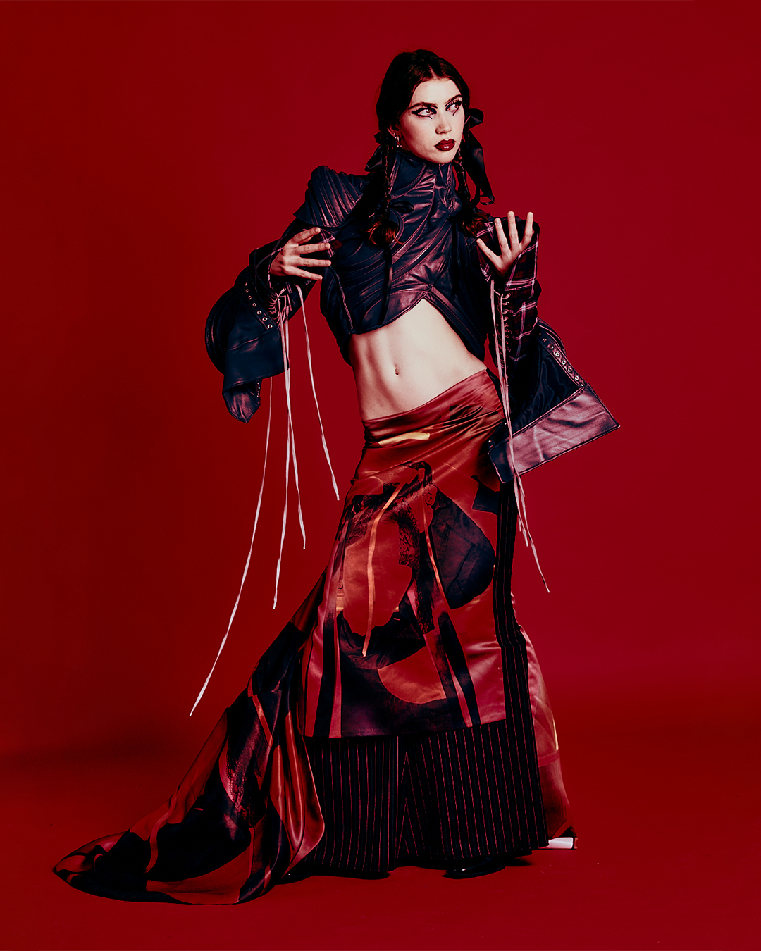 A woman with dark hair, bold makeup, and a piercing stare, dressed in a black and red fashion outfit, poses confidently against a red background.