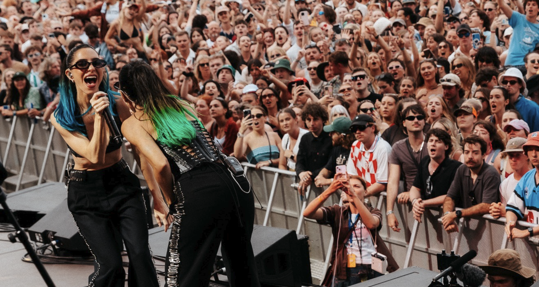 Two performers singing on stage at a concert with a large crowd cheering and taking photos.