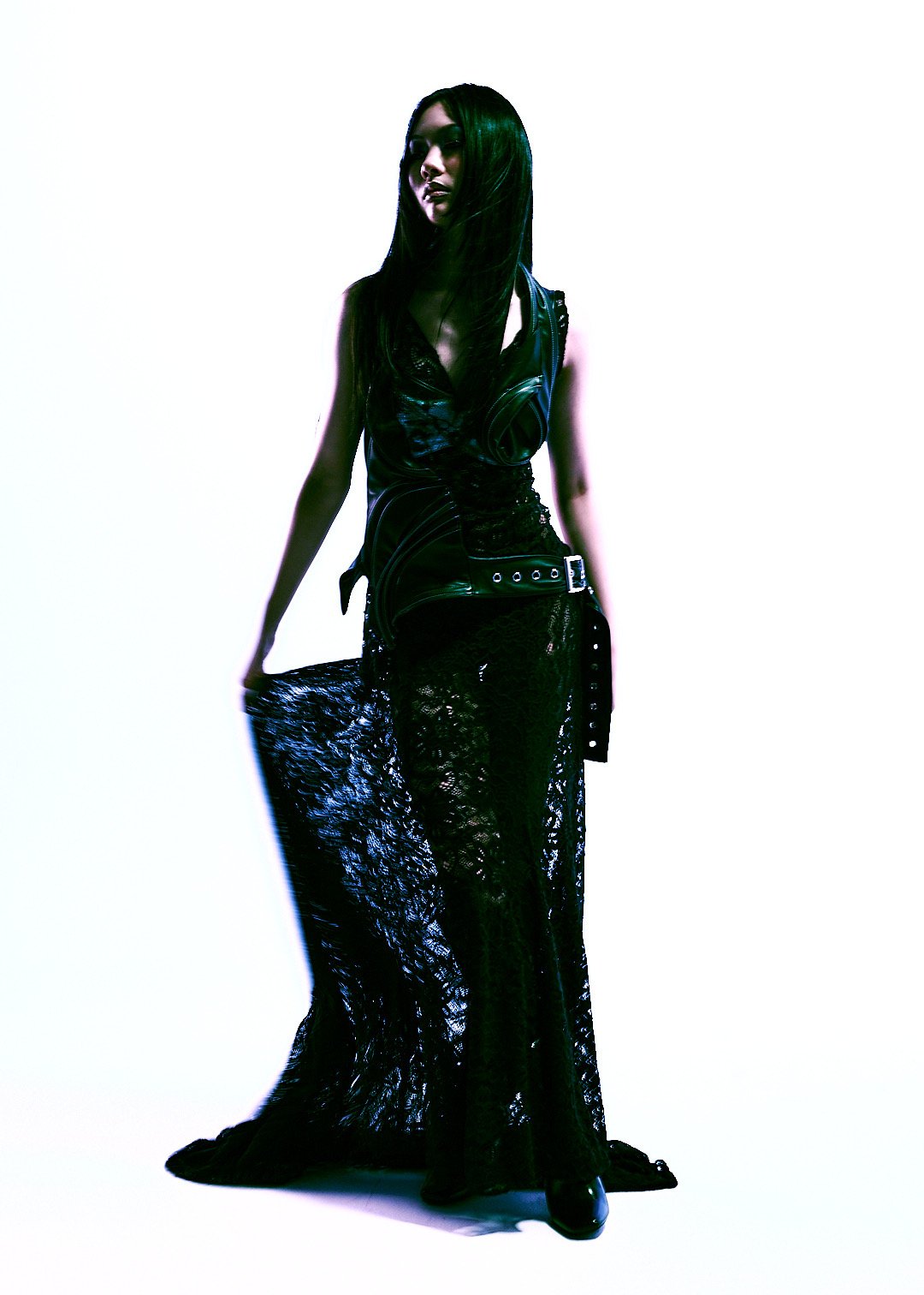 Woman in black leather outfit and lace skirt standing against a plain background with minimal lighting.