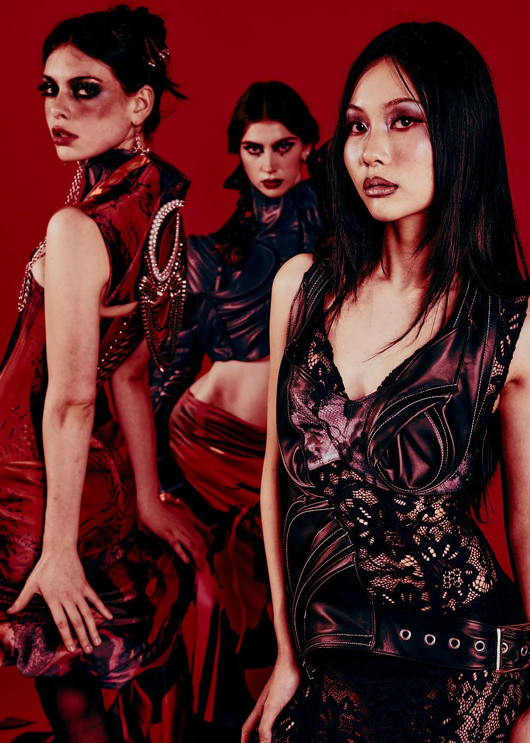 Three women posing against a red background, wearing dark, elaborate, and edgy fashion outfits with makeup.