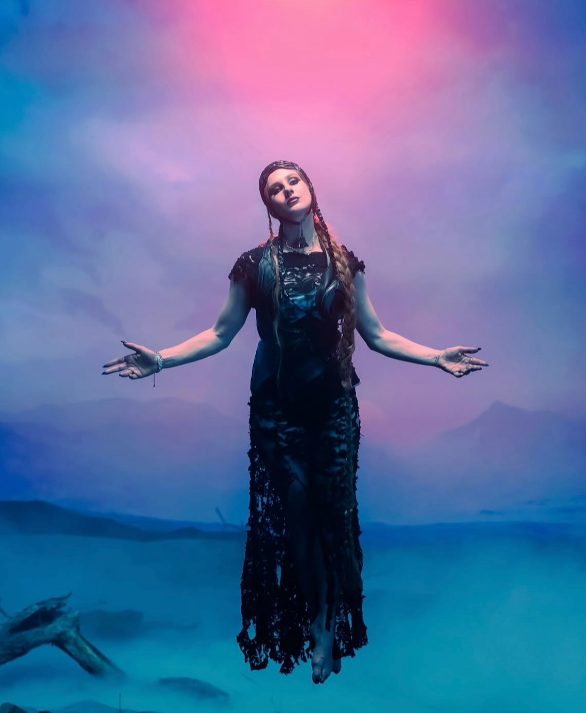 A woman with long braided hair stands in a foggy landscape with a colorful sky, wearing a black lace dress.