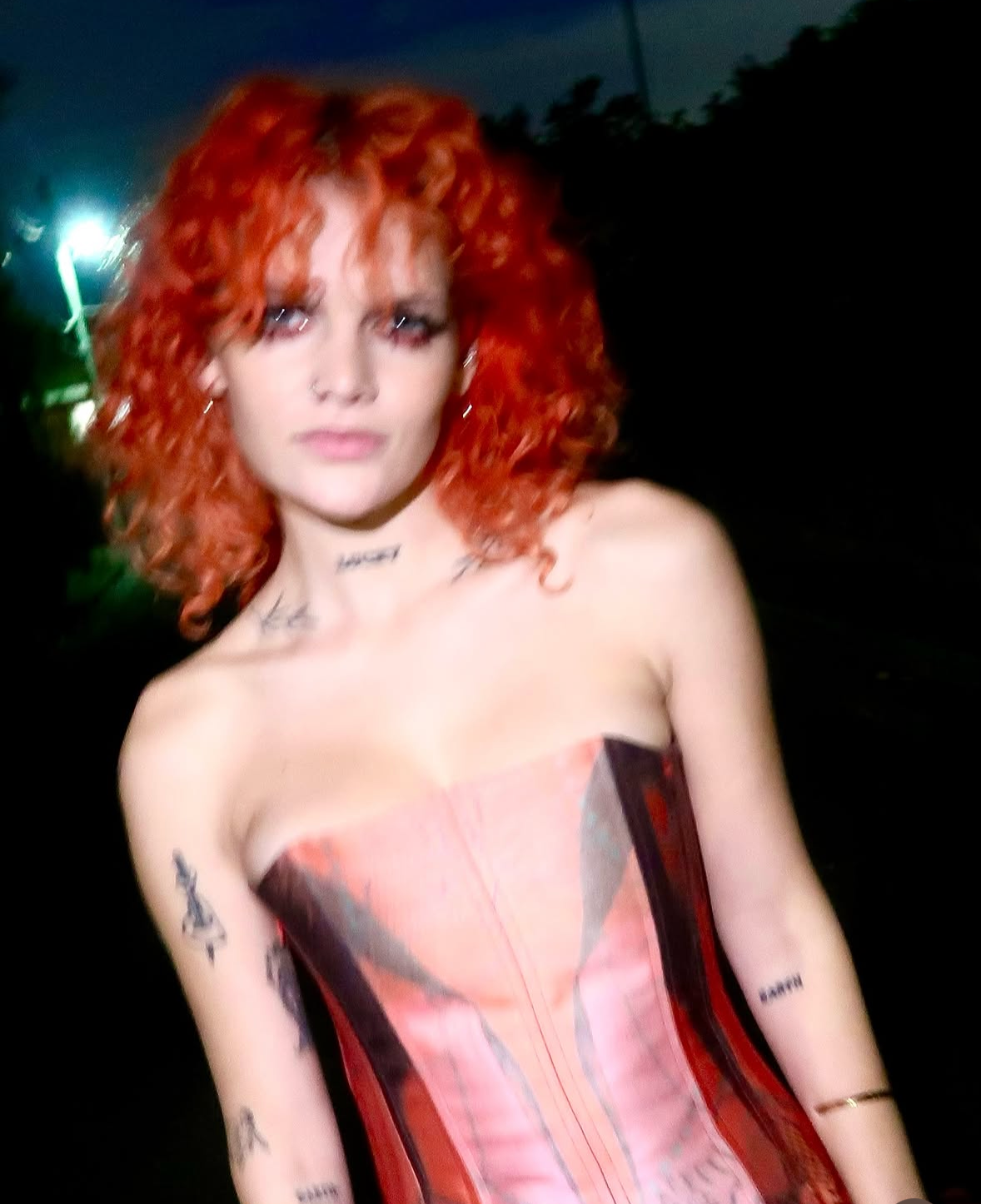 A woman with curly red hair and tattoos on her arms and neck, wearing a strapless dress or top, standing outdoors at night with a dark background and faint street lights.