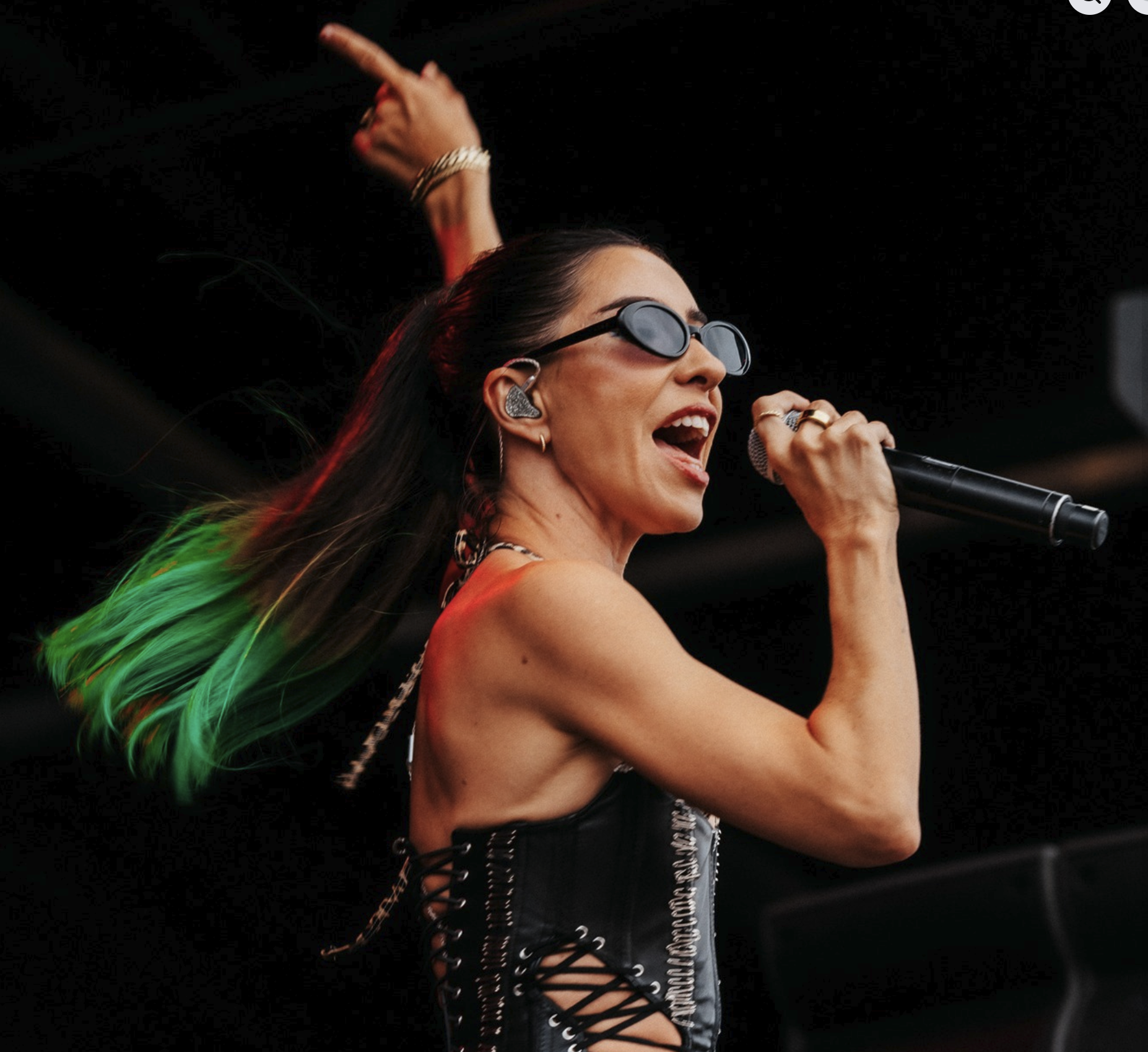 A woman singing into a microphone on stage with long brown hair, wearing sunglasses, earrings, and a black dress with side lace-up details, with multicolored hair that includes green at the tips.