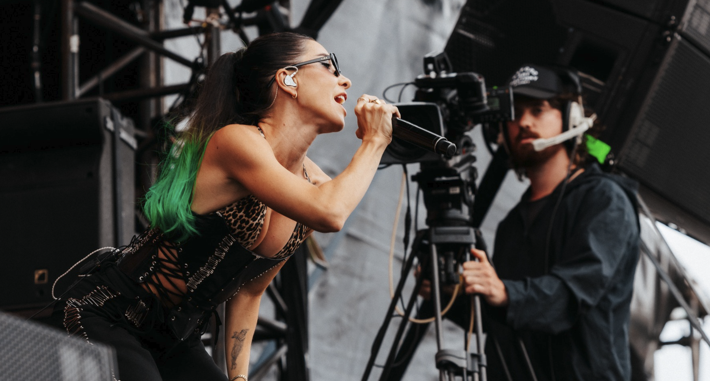 A woman with long dark hair, sunglasses, and a leopard print outfit is singing into a microphone on a stage, with a cameraman recording her in the background.