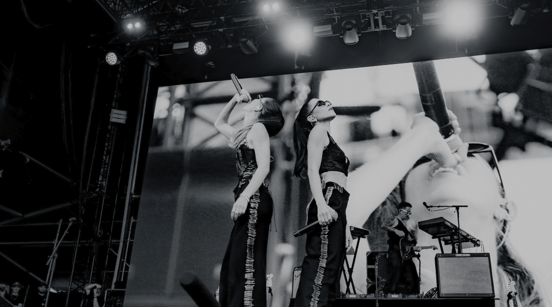 Two female singers performing on stage with microphones, with a large screen showing a close-up of one of the singers in the background. There are musicians and equipment on stage under bright stage lights.