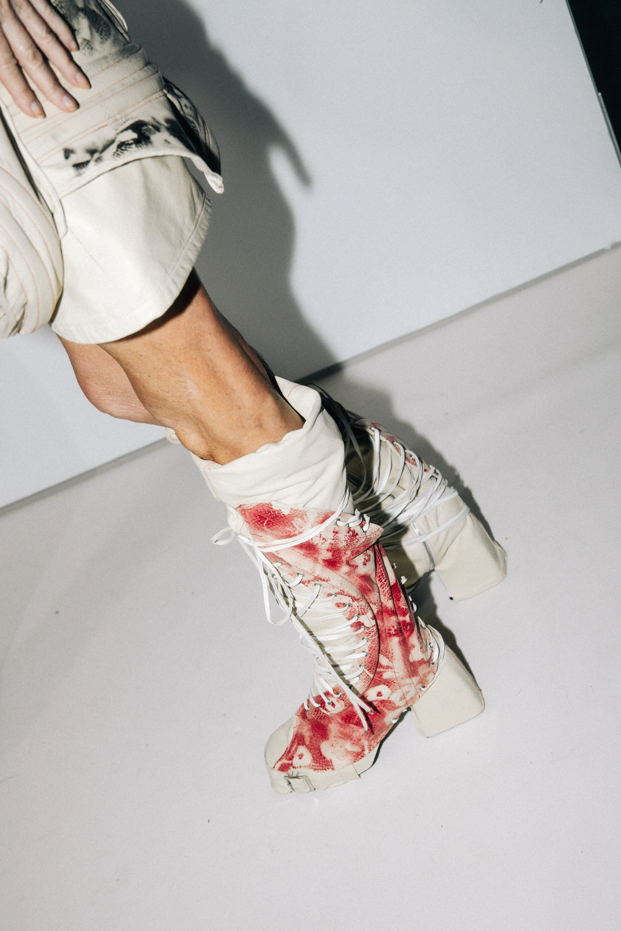 Close-up of a person wearing cream-colored high-top sneakers with red paint splatters, cream-colored cargo shorts, and a beige t-shirt, standing on a white floor.