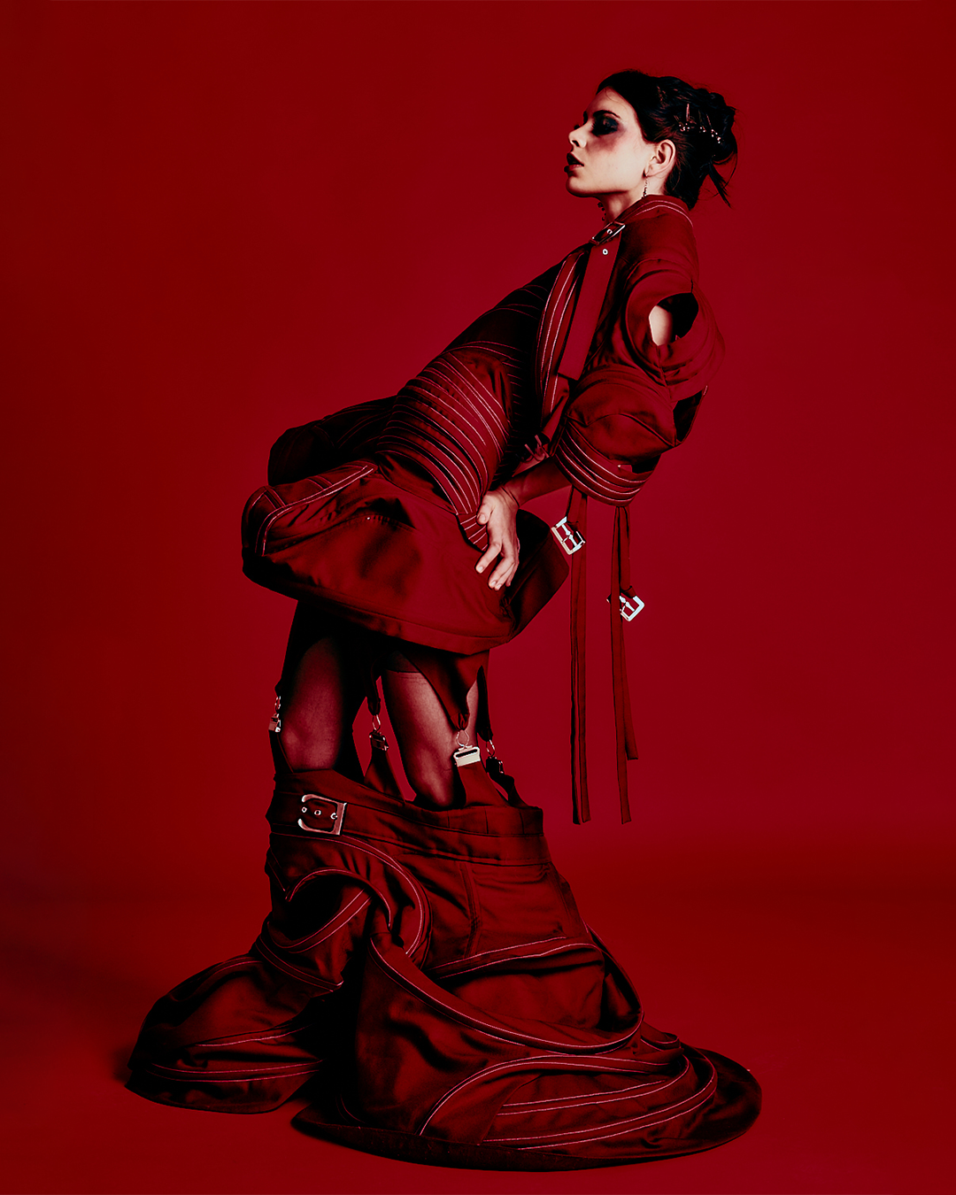 A woman dressed in red, wearing avant-garde fashion with layered fabrics, straps, and zippers, standing against a red background.