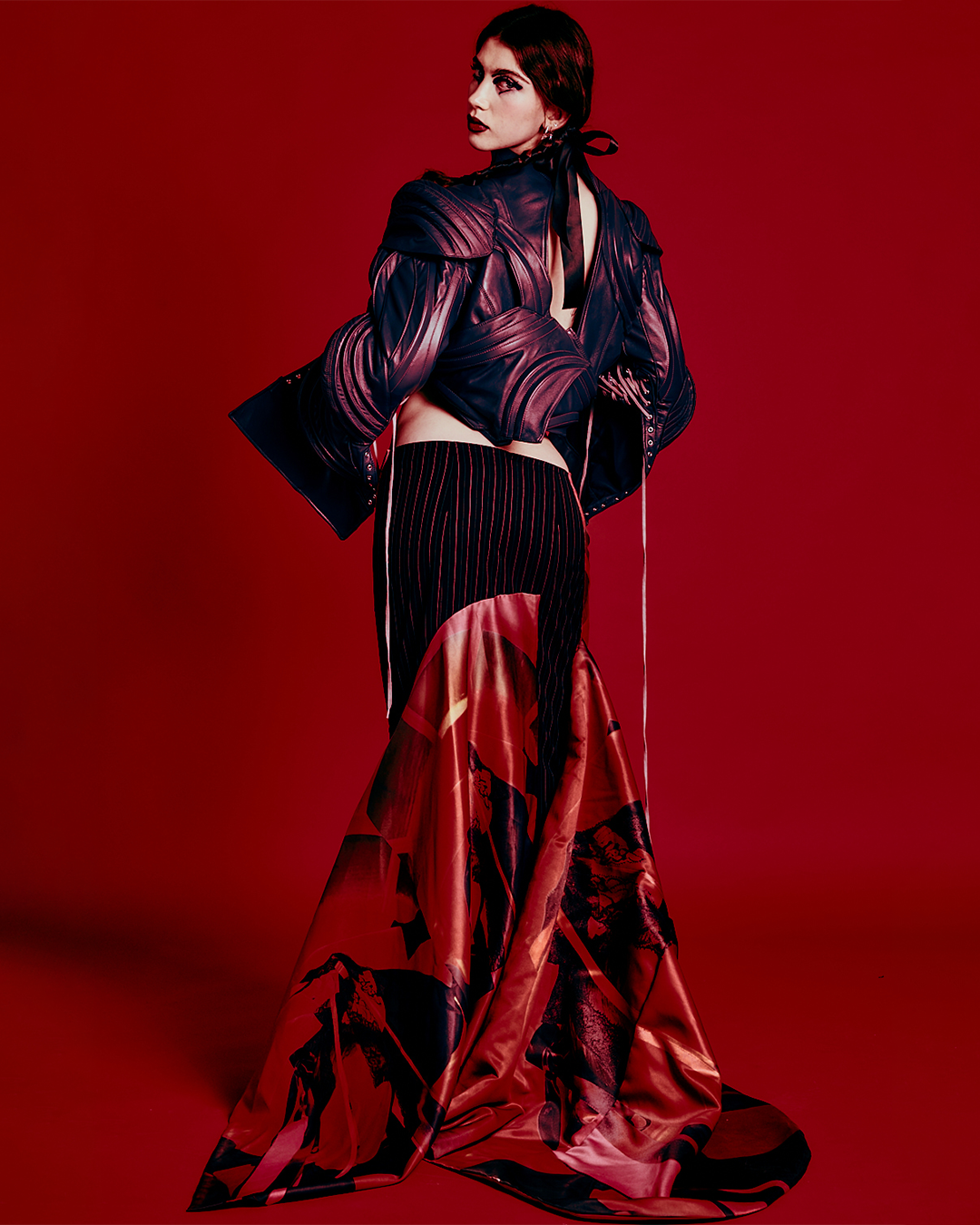 A woman with dark hair and makeup wearing a shiny, dark, leather-like jacket over a dark top and a long, patterned skirt with red, black, and cream colors, standing against a red background.