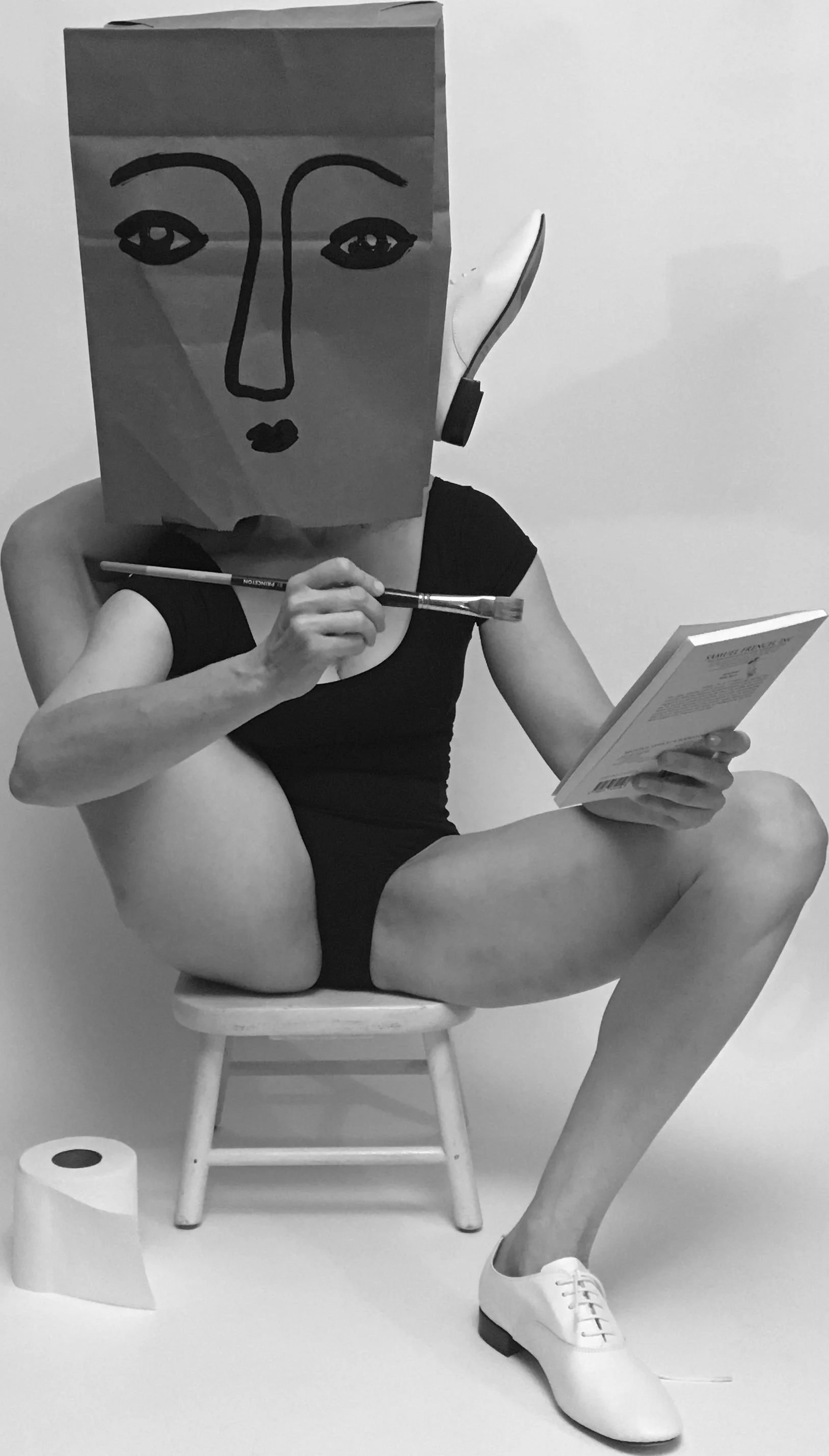 A person sitting on a small wooden stool, wearing a black sleeveless top and white shoes, with a paper bag on their head featuring a simple face drawing. They are holding a paintbrush in one hand and a book in the other, with a roll of toilet paper on the floor in front of them.