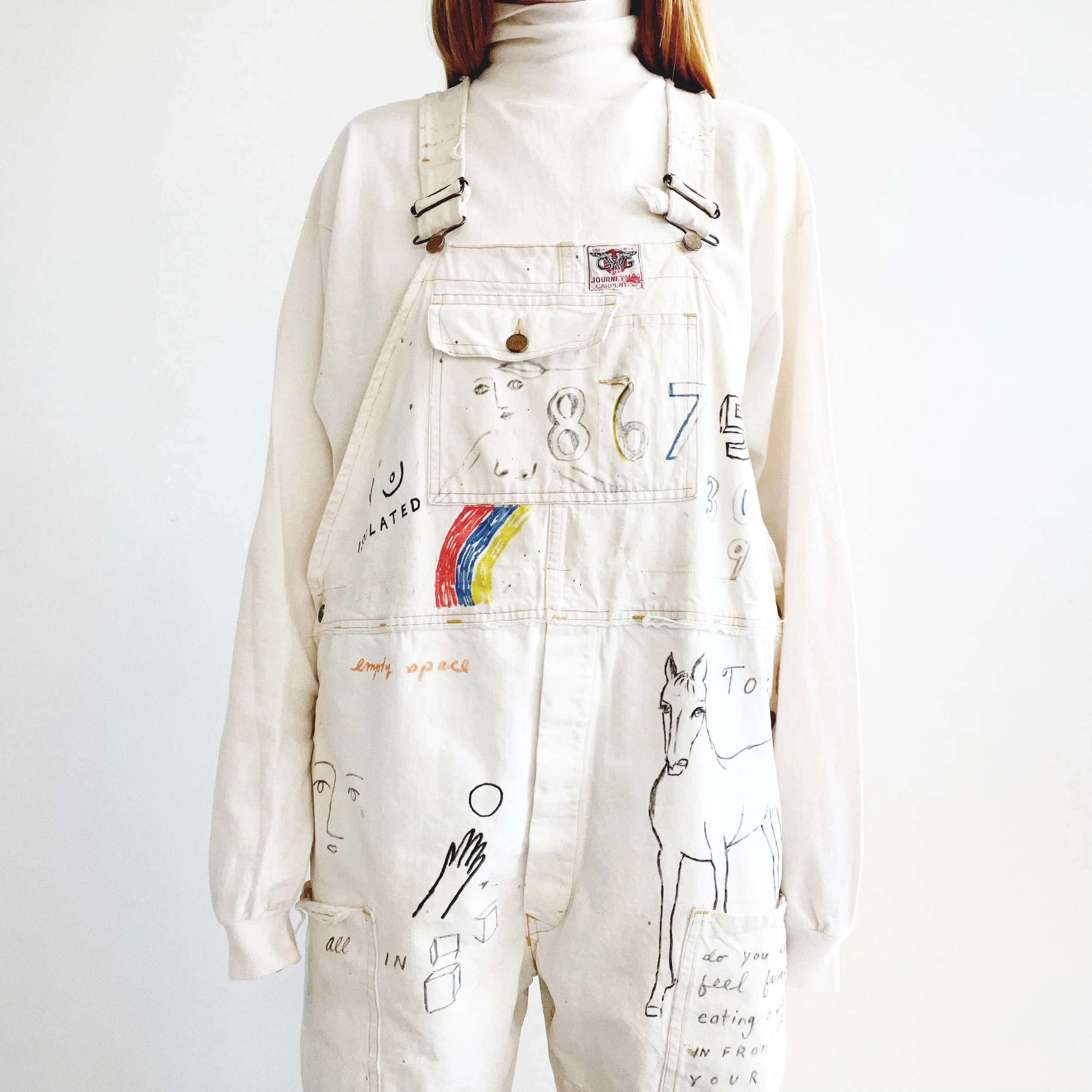 A person wearing a white overall shirt with colorful drawings, handwritten words, and doodles. The drawings include a rainbow, a face, and animals, while the words include phrases like 'empty space' and 'feeding'. The person has long hair and stands against a plain white background.