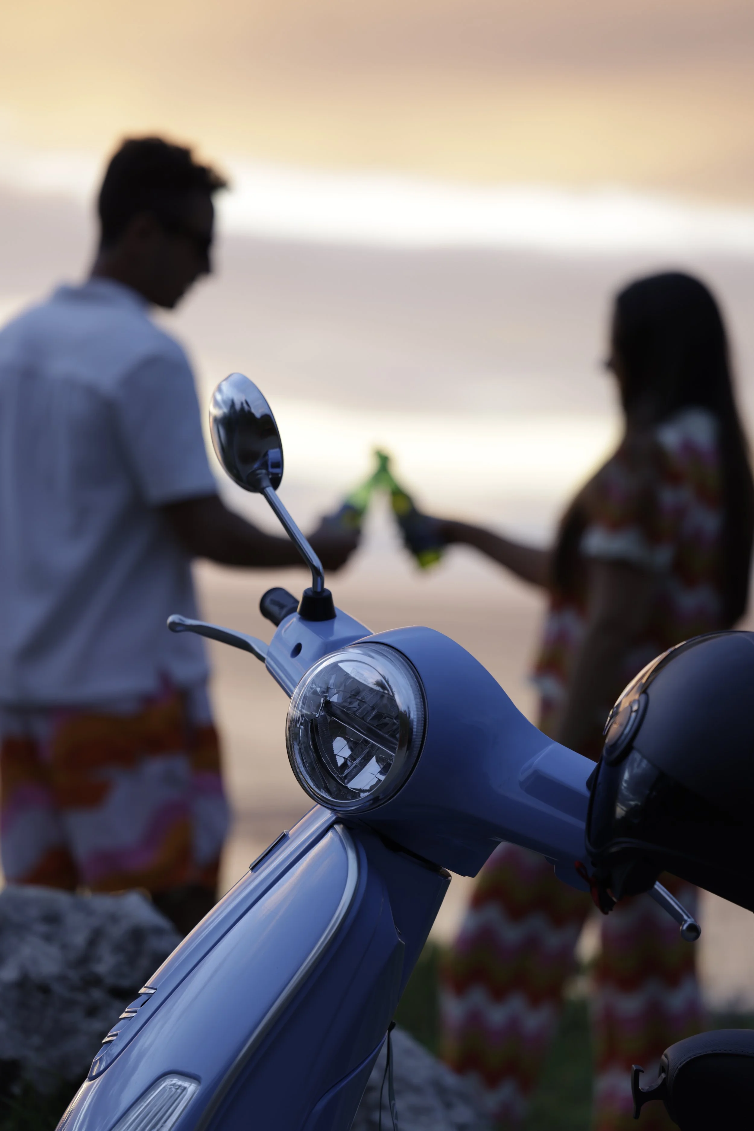 Close-up of a blue scooter in the foreground, with two people holding drinks and engaging in conversation in the blurred background during sunset.
