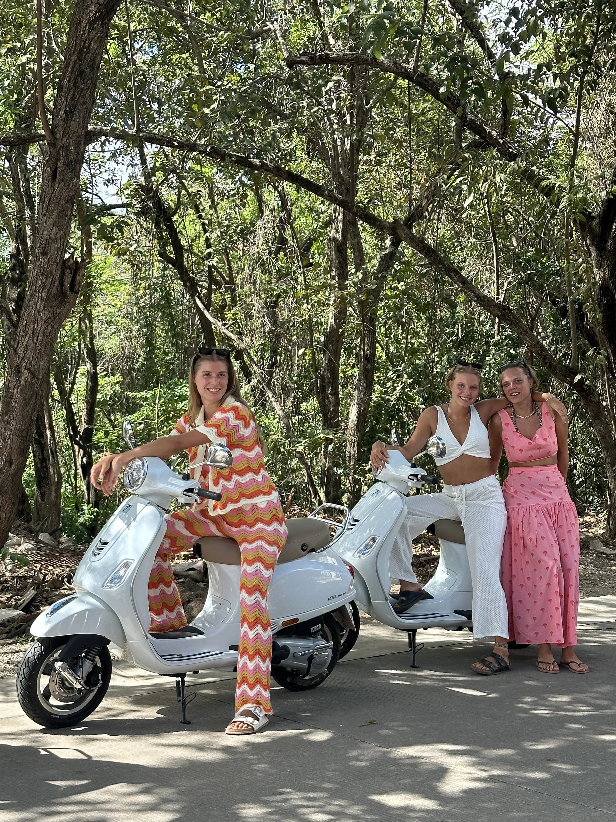 Three young women with blonde hair and casual summer outfits posing with white scooters in a wooded outdoor setting.