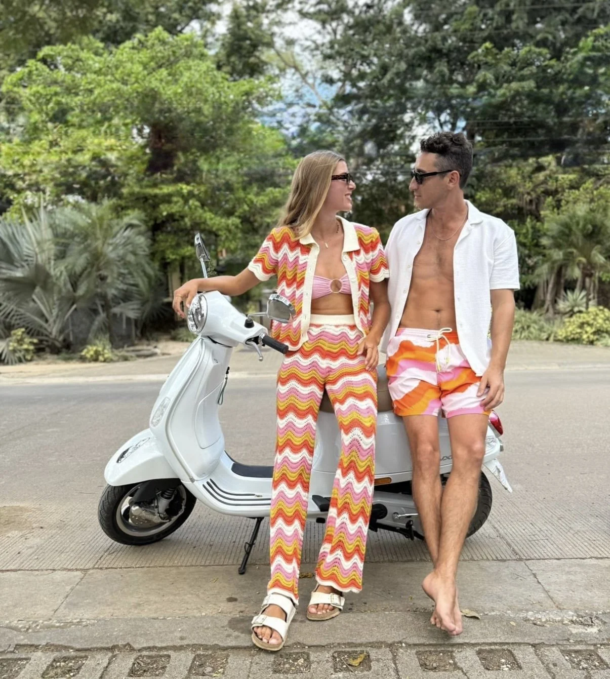 A man and woman in colorful beach attire standing next to a white scooter on a street with greenery. The woman wears a zigzag patterned top and pants with pink, orange, yellow, and white colors, and the man wears swim trunks and an open white shirt, both wearing sunglasses.
