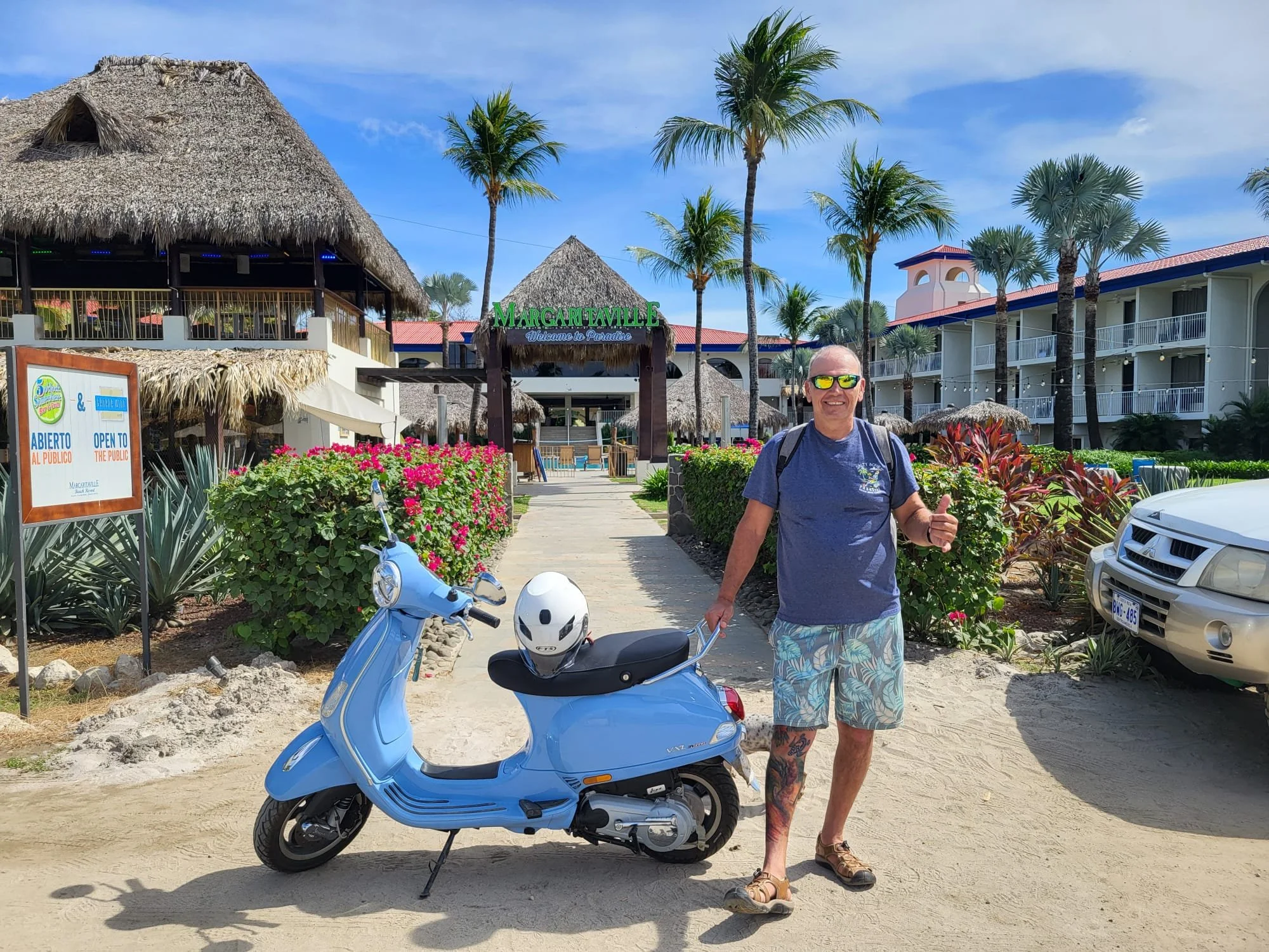 A man wearing sunglasses, a blue t-shirt, and tropical shorts standing next to a blue scooter with a white helmet on the seat, in front of a hotel resort named Margaritaville with palm trees and a clear sky.