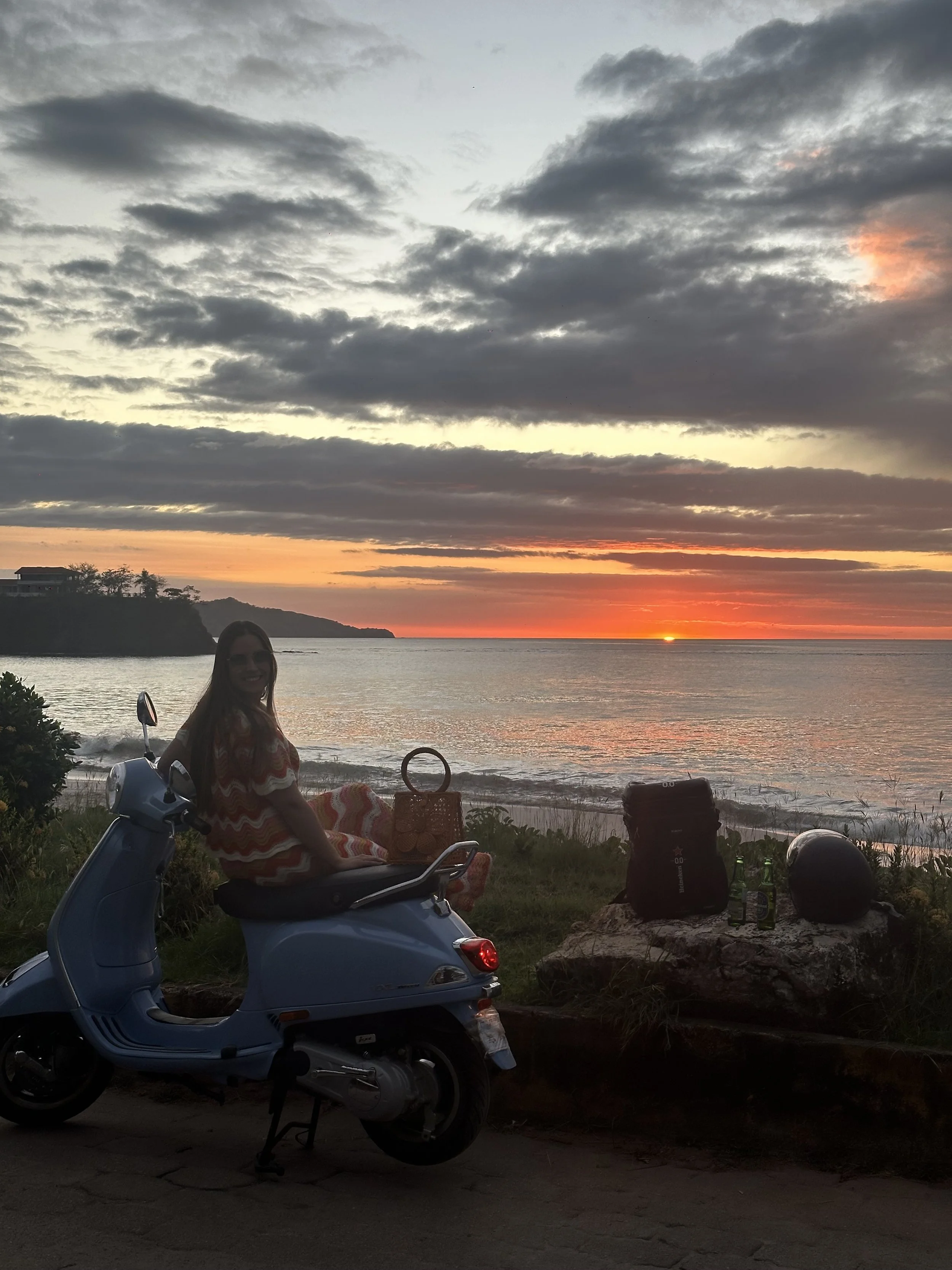 A woman sitting on a blue scooter parked by the beach at sunset, with a sunset sky and mountains in the background. There are bags, bottles, and a helmet on the ground nearby.