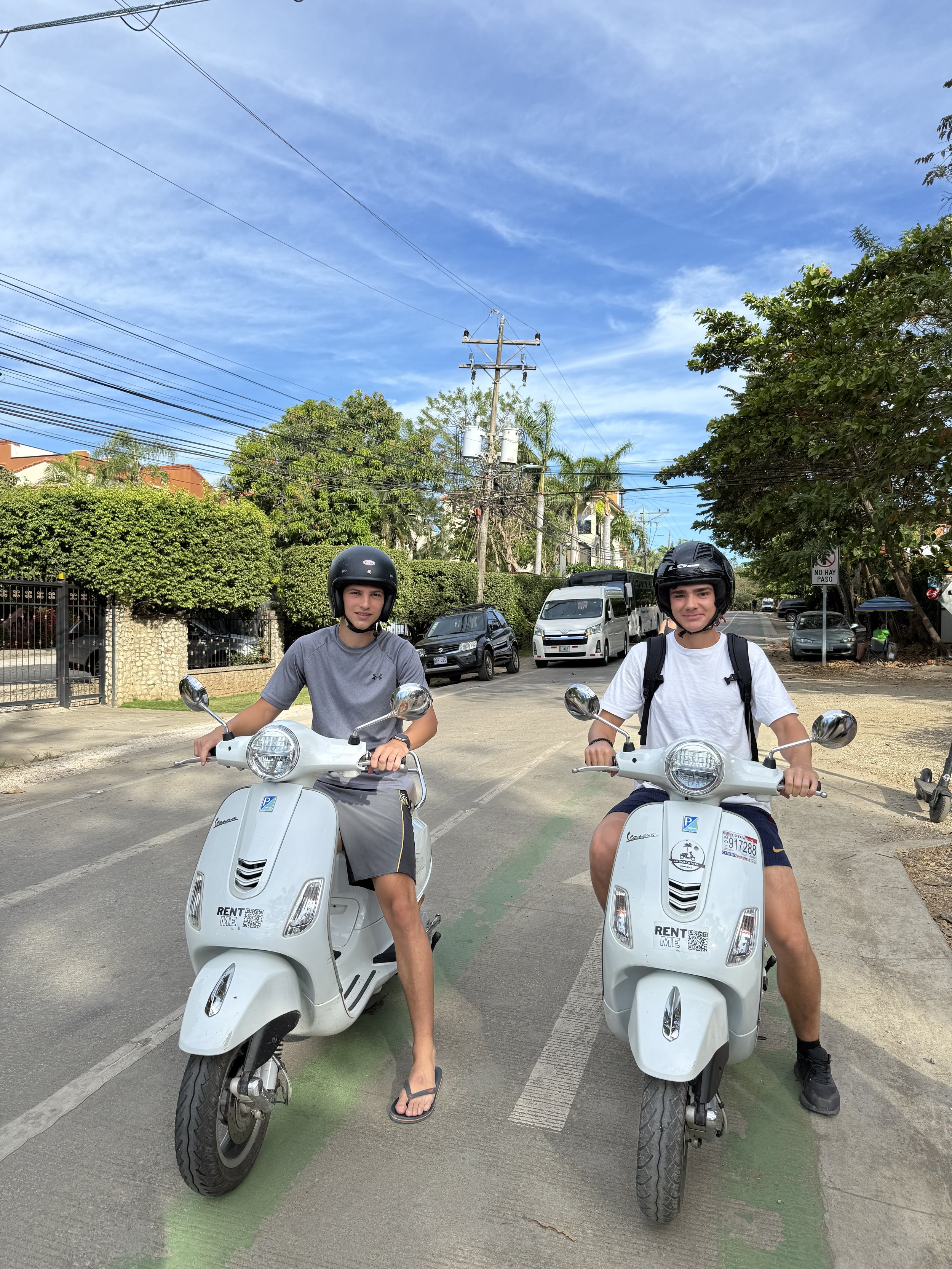 Two young men wearing helmets sitting on white scooters in a street with cars and green trees in the background on a sunny day.