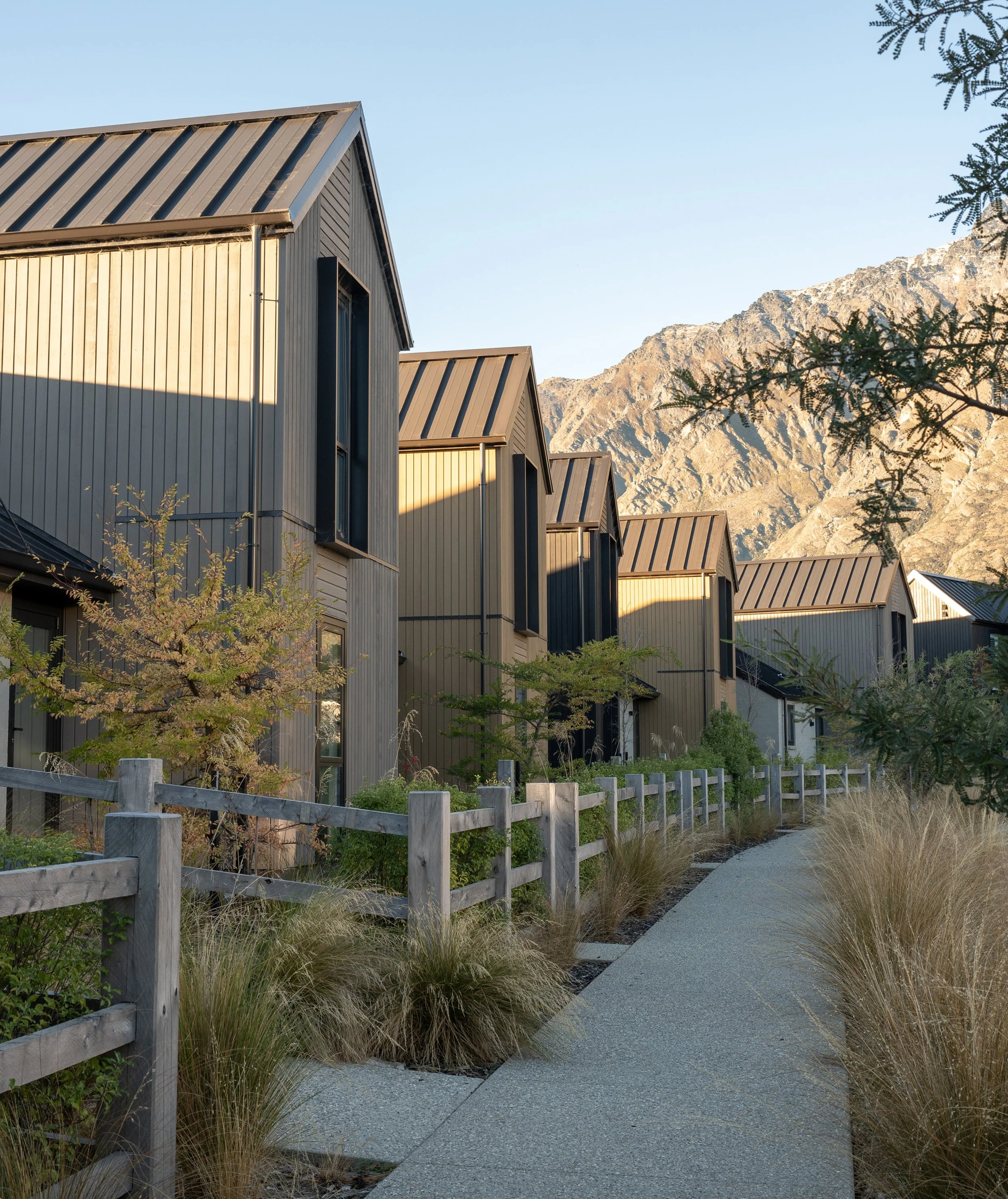 Residential neighborhood with modern houses and mountains in the background, sunlight illuminating the scene, modern gray fences, plants, and a gravel pathway.