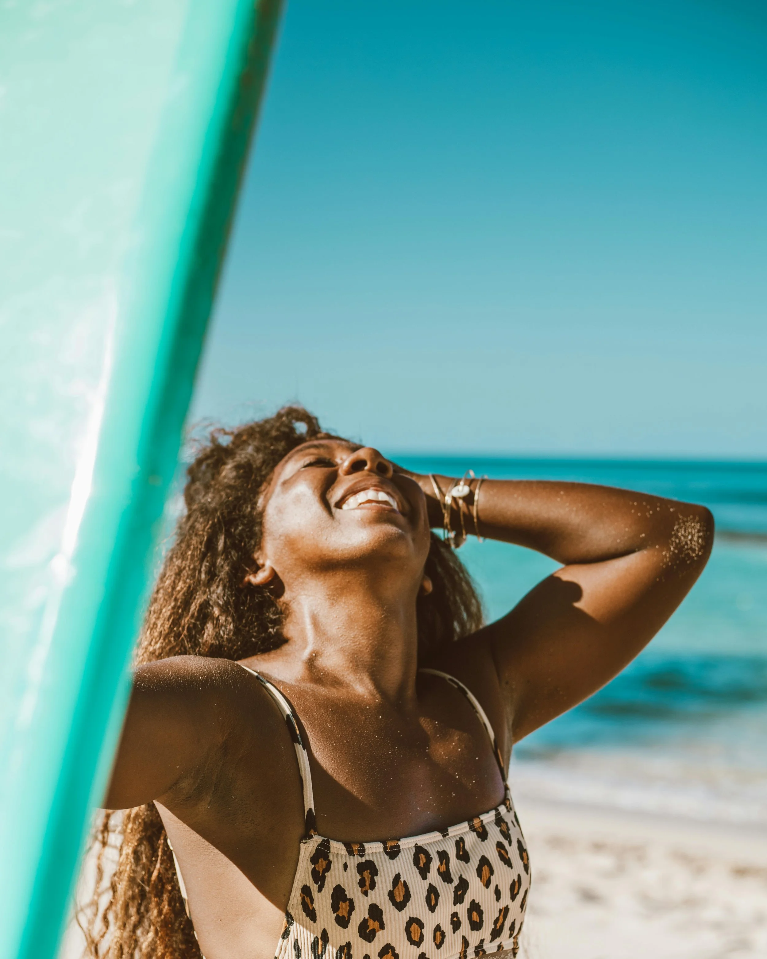 Woman smiling on a beach with a surfboard beside her.