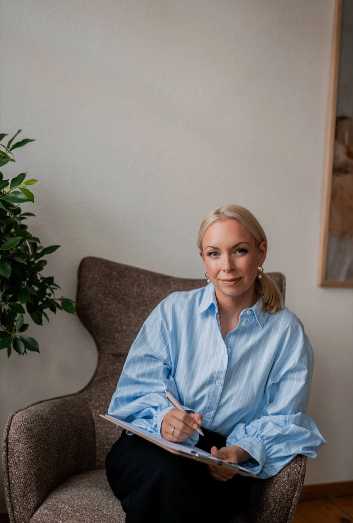 Eine blonde Frau sitzt in einem braunen Sessel, trägt eine hellblaue, gestreifte Bluse, hält einen Notizblock und einen Stift und lächelt.