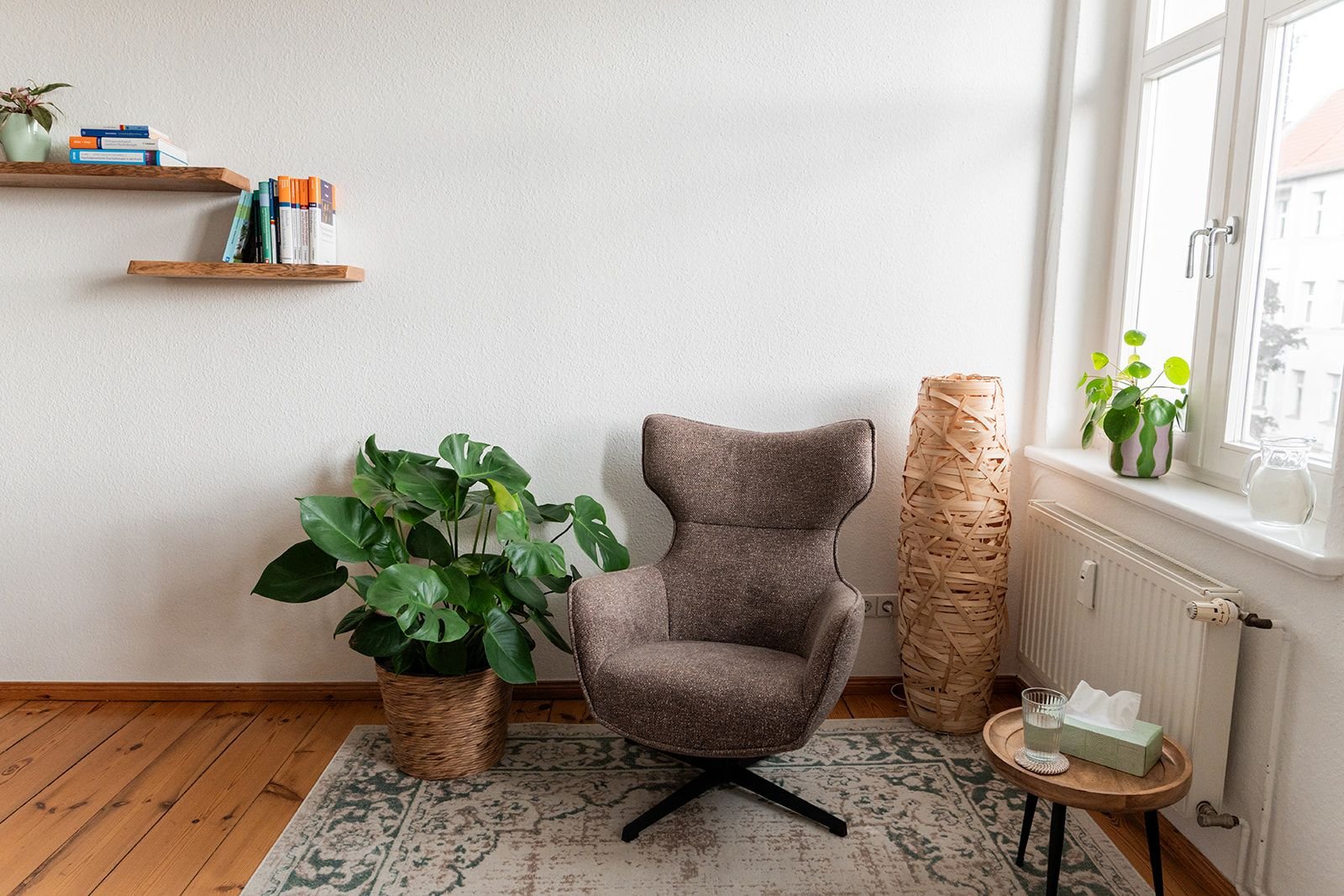 Gemütliches Wohnzimmer mit einem braunen Sessel, einer großen Zimmerpflanze in einem Korb, einem kleinen Beistelltisch mit Wasser, einem Packung Taschentücher und einem Glas, einem Fenster mit Blick nach draußen, einer dekorativen Papierlampe und einer Wand mit zwei Holzregalen, die Bücher tragen.