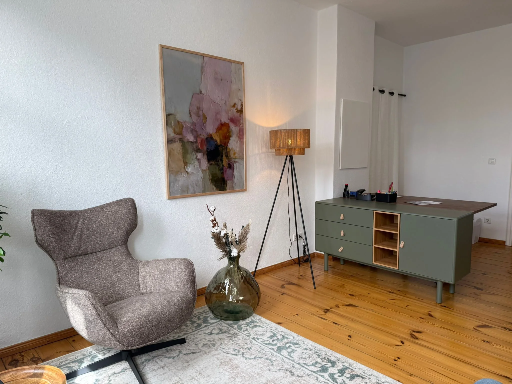 Ein Wohnzimmer mit einem grauen Sessel, einer großen Vase mit Pflanzen, einem Wandbild, einem Stehlampe, einem grünen Sideboard und weißen Vorhängen.