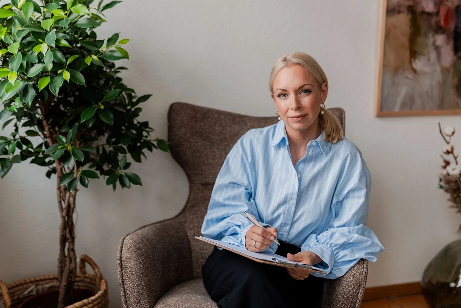 Eine blonde Frau sitzt auf einem braunen Sessel, trägt eine hellblaue gestreifte Bluse und hält einen Dokumentenordner mit einem Stift in der Hand, um ihre Notizen zu machen.