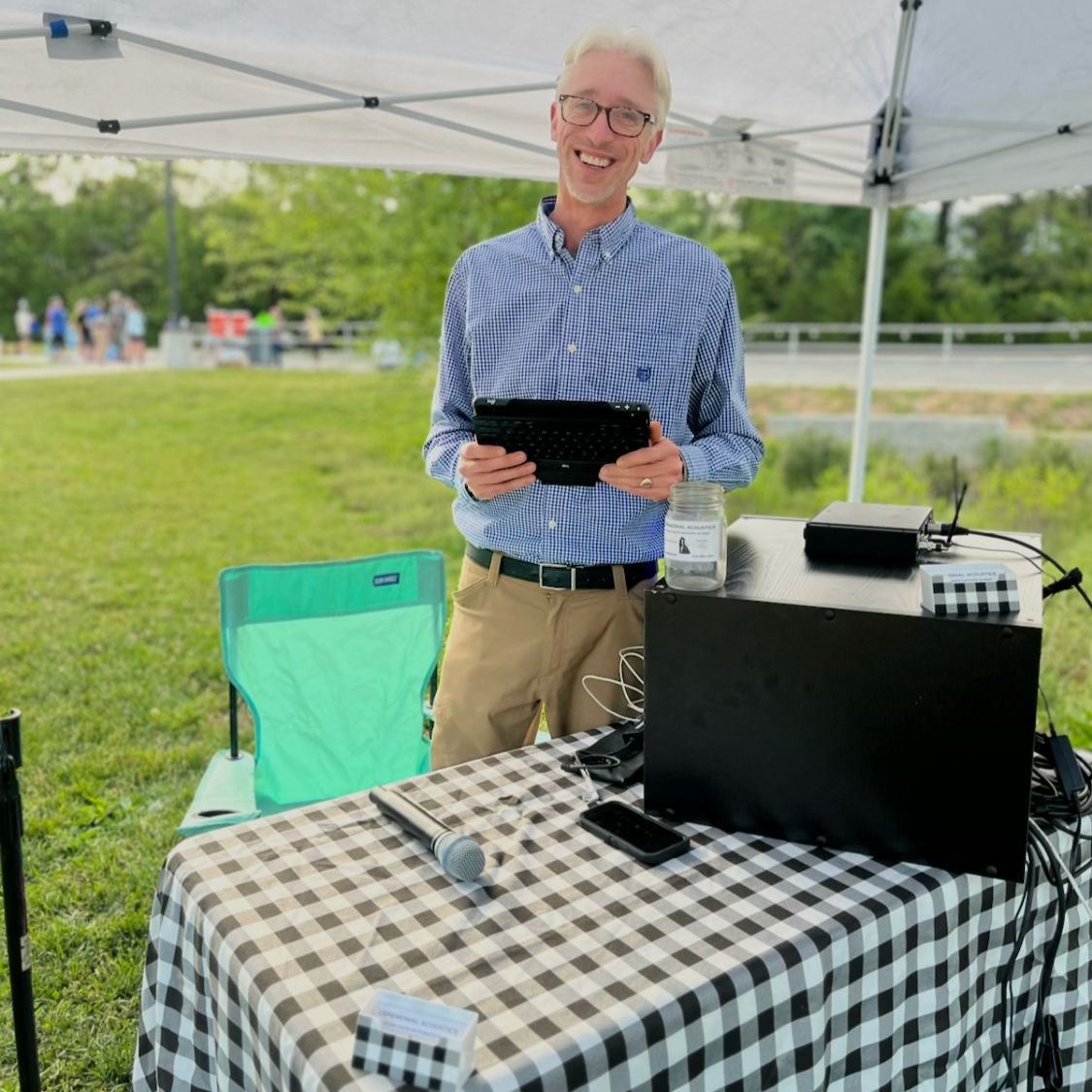 A smiling man standing under a white canopy at an outdoor event, holding a tablet. There is a table with a microphone, a smartphone, and other electronic equipment, and a green and blue chair nearby.