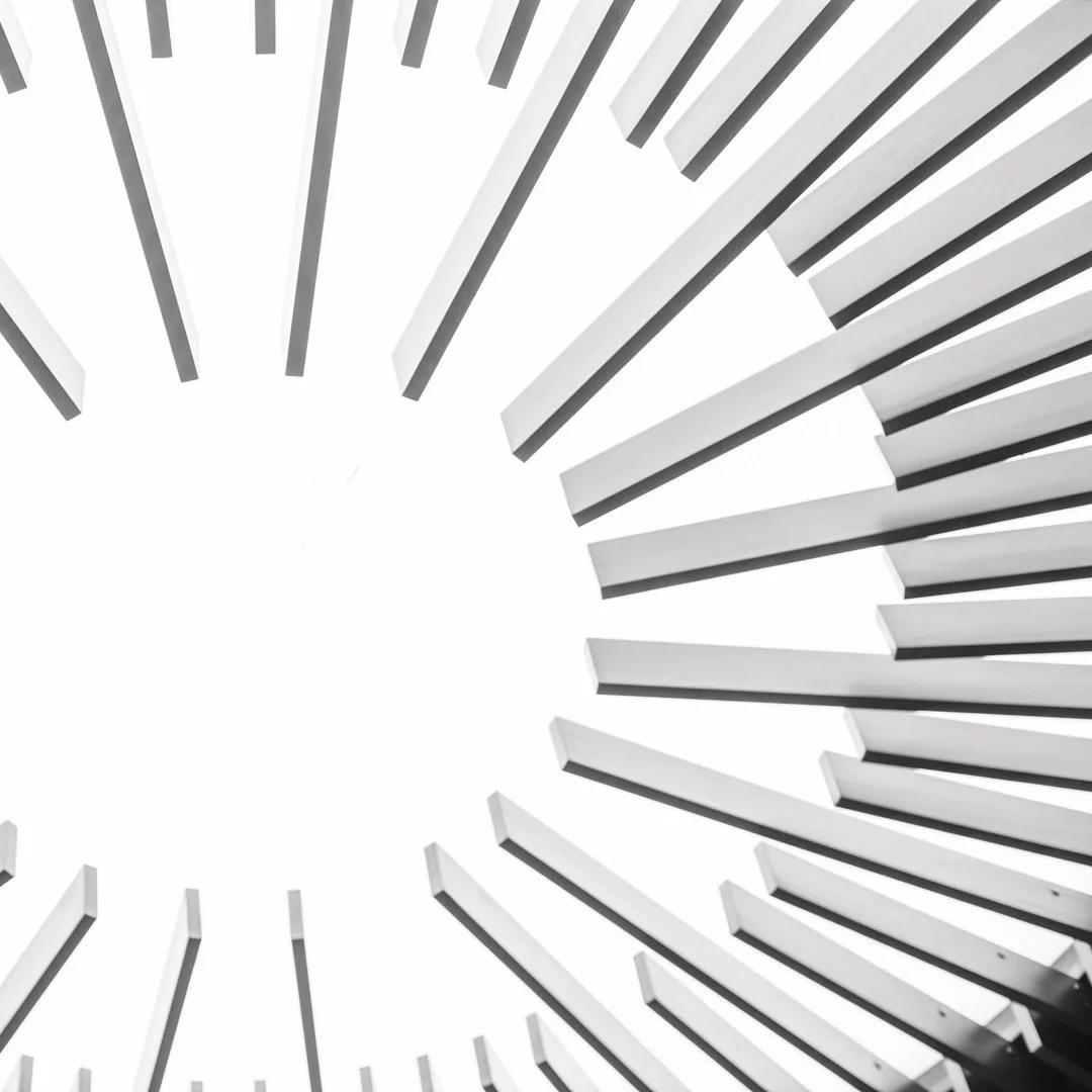 A modern architectural structure with white elongated beams arranged in a circular pattern, viewed from below against a white sky.
