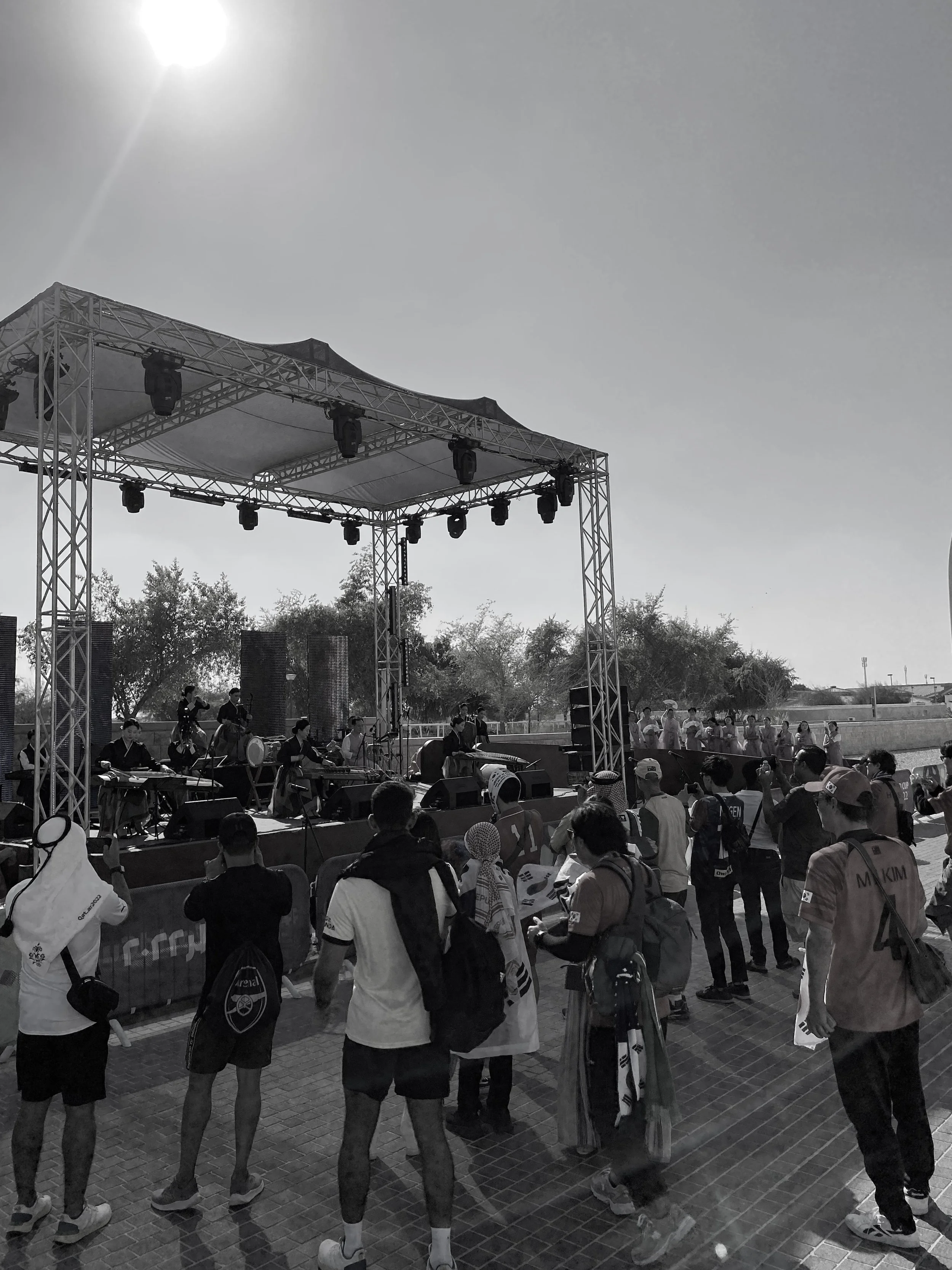 A crowd of people gathers around a stage set outdoors, with trees and a clear sky in the background. Music equipment and performers are on the stage, and some audience members are standing and watching.
FIFA Men's World Cup Qatar 2022. Doha, Qatar