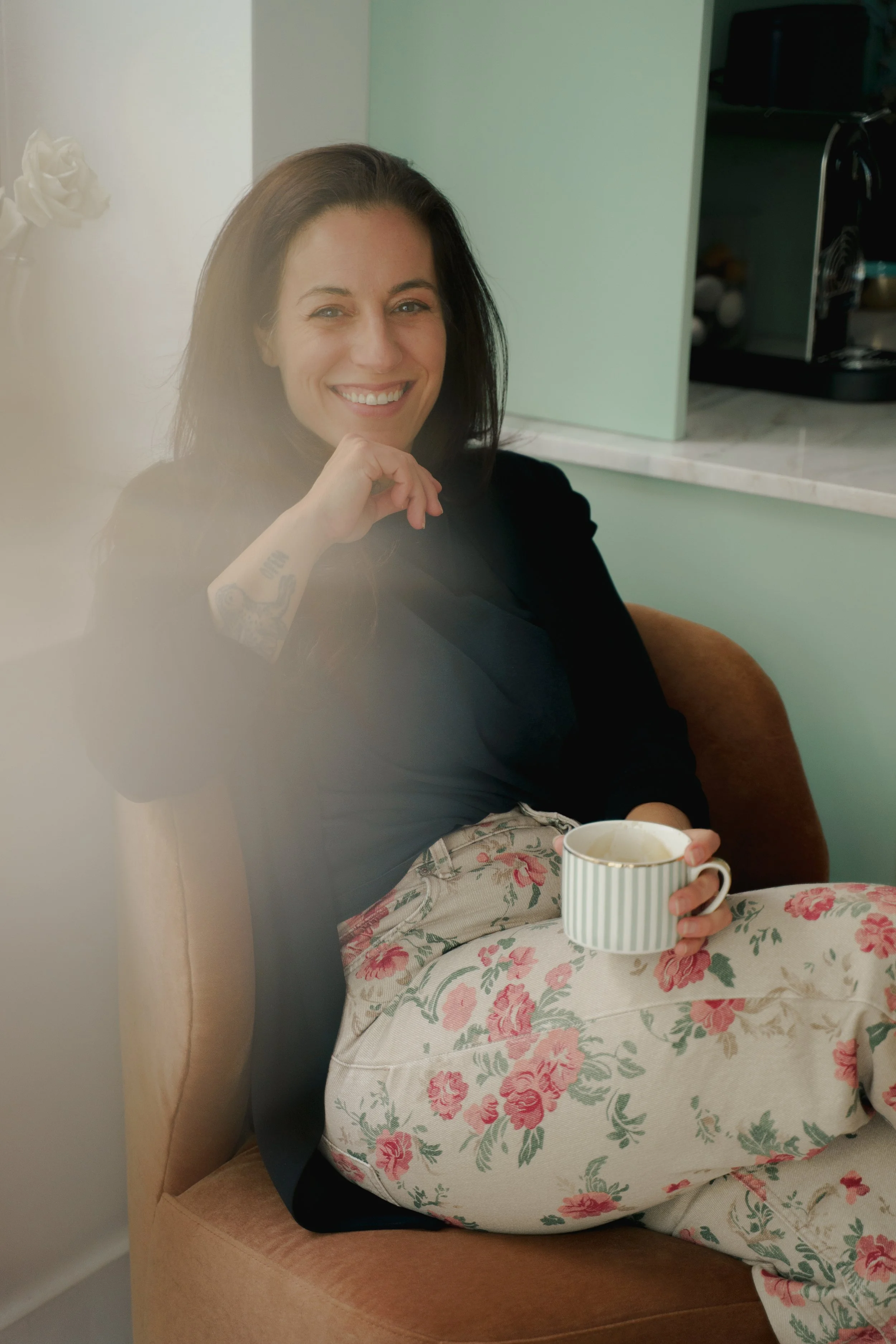 A woman with dark hair smiling holds a mug with a striped pattern, sitting on a floral covered table, while leaning on a cushioned armchair in a kitchen.