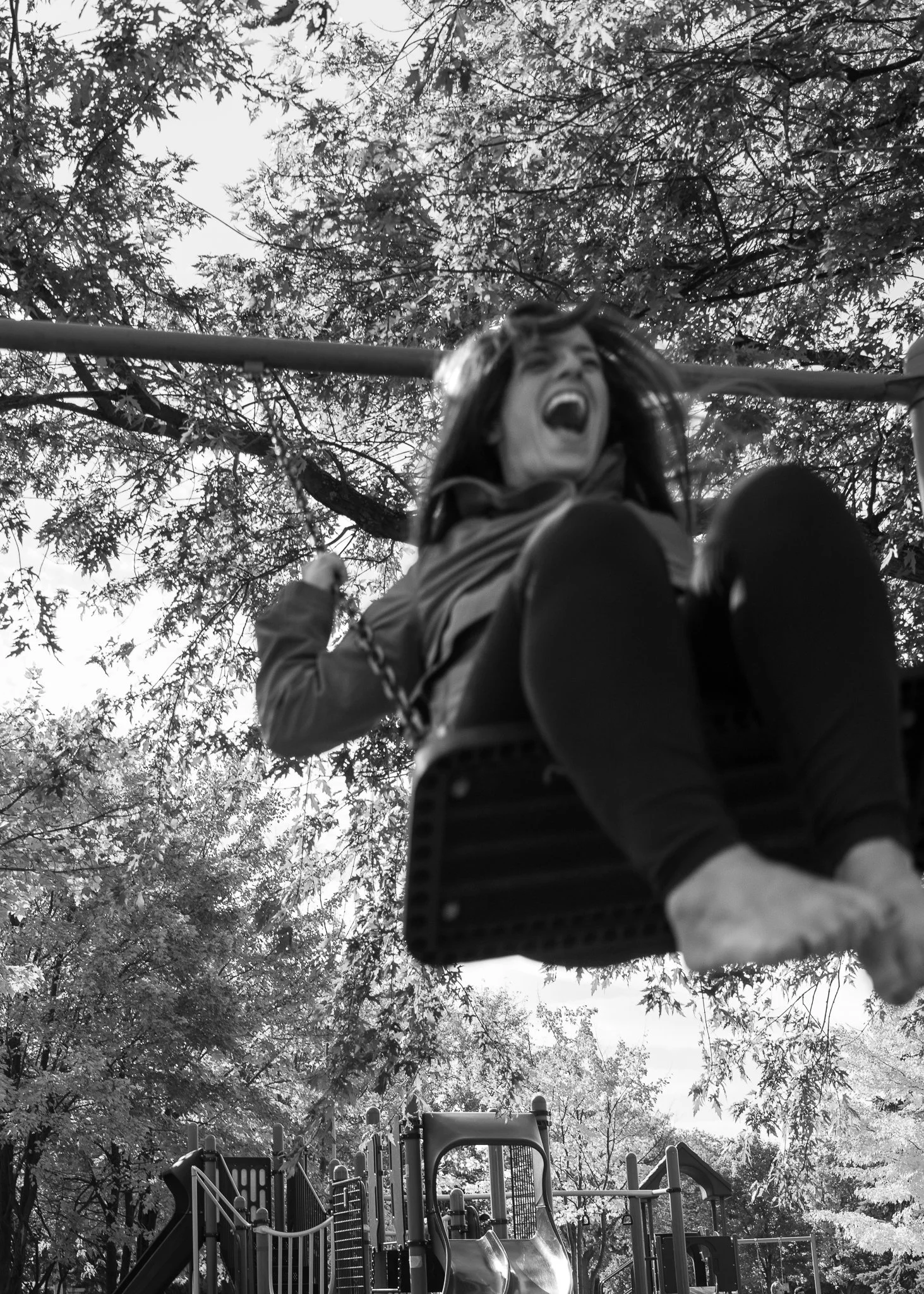 A woman with long hair on a playground swing, smiling and laughing, with a canopy of trees in the background.