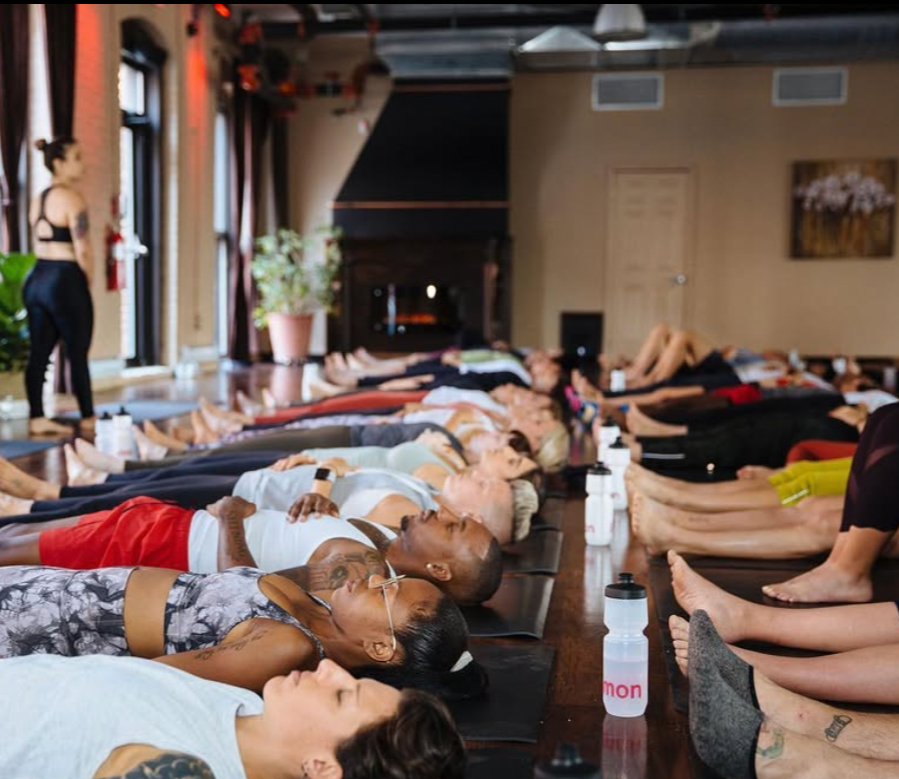 Group of people participating in a yoga class in a studio, lying on mats with feet toward the instructor, who is standing at the front of the room.