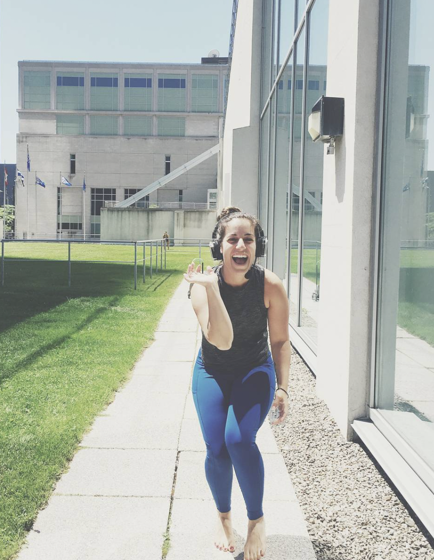 Young woman with headphones smiling and walking barefoot outside on a sidewalk near a modern building.