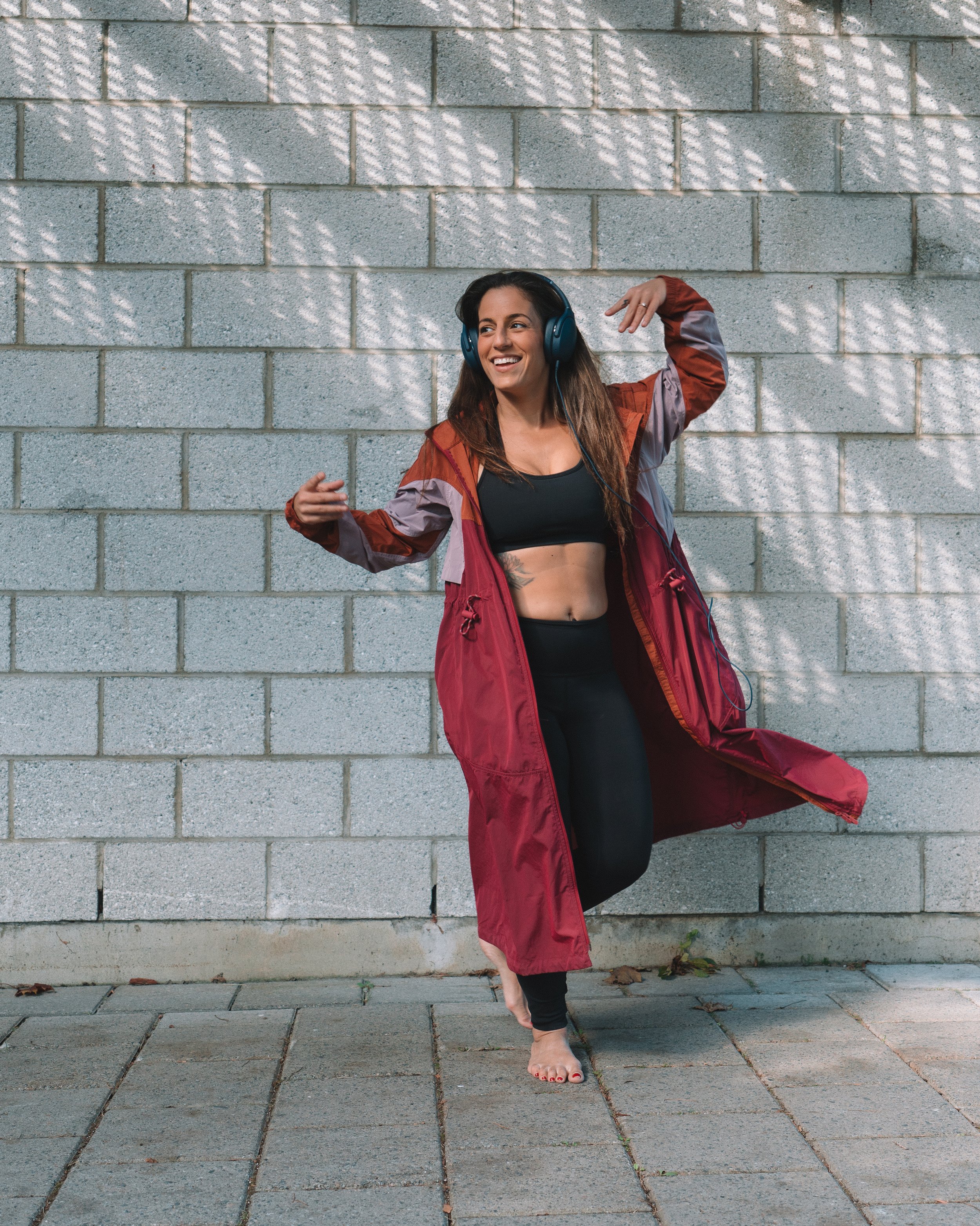 A woman with brown hair wearing headphones, a black sports bra, and a red jacket standing on one leg with her arms in dance pose against a brick wall.