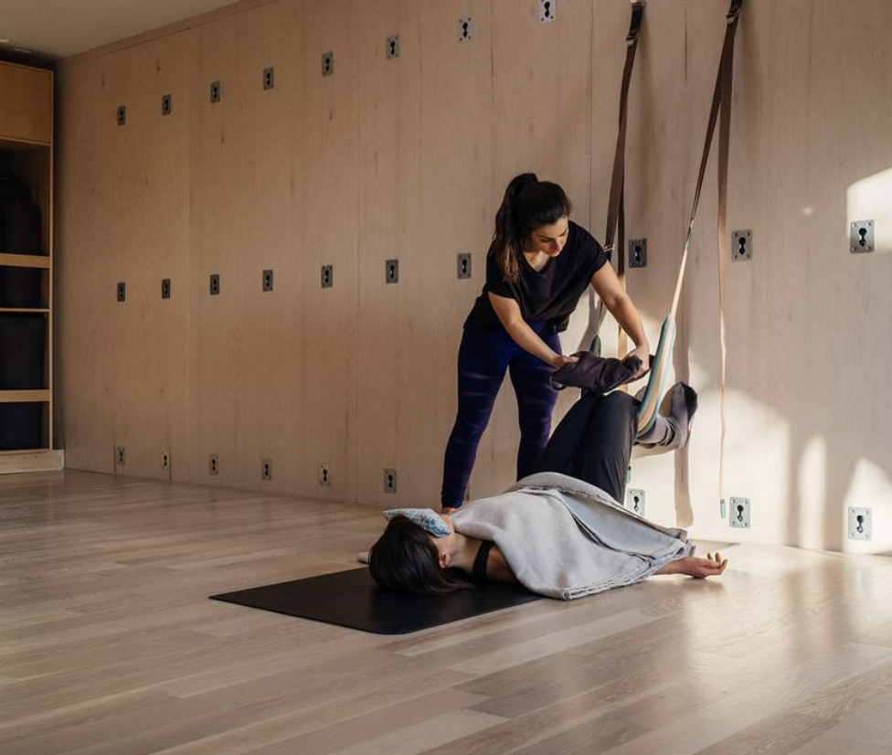 A woman lying on a yoga mat with an eye pillow over her eyes, receiving assistance from an instructor in a yoga or meditation setting, with sunlight streaming in.