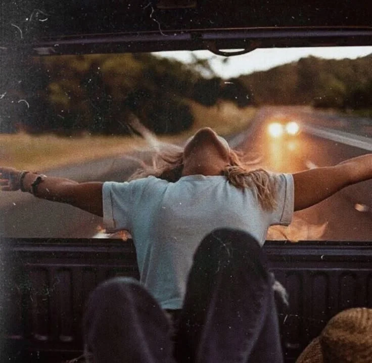 A woman in a white t-shirt sitting in the back of a vehicle with her arms stretched out and head tilted back, enjoying the view of the road and sunset through the windshield.